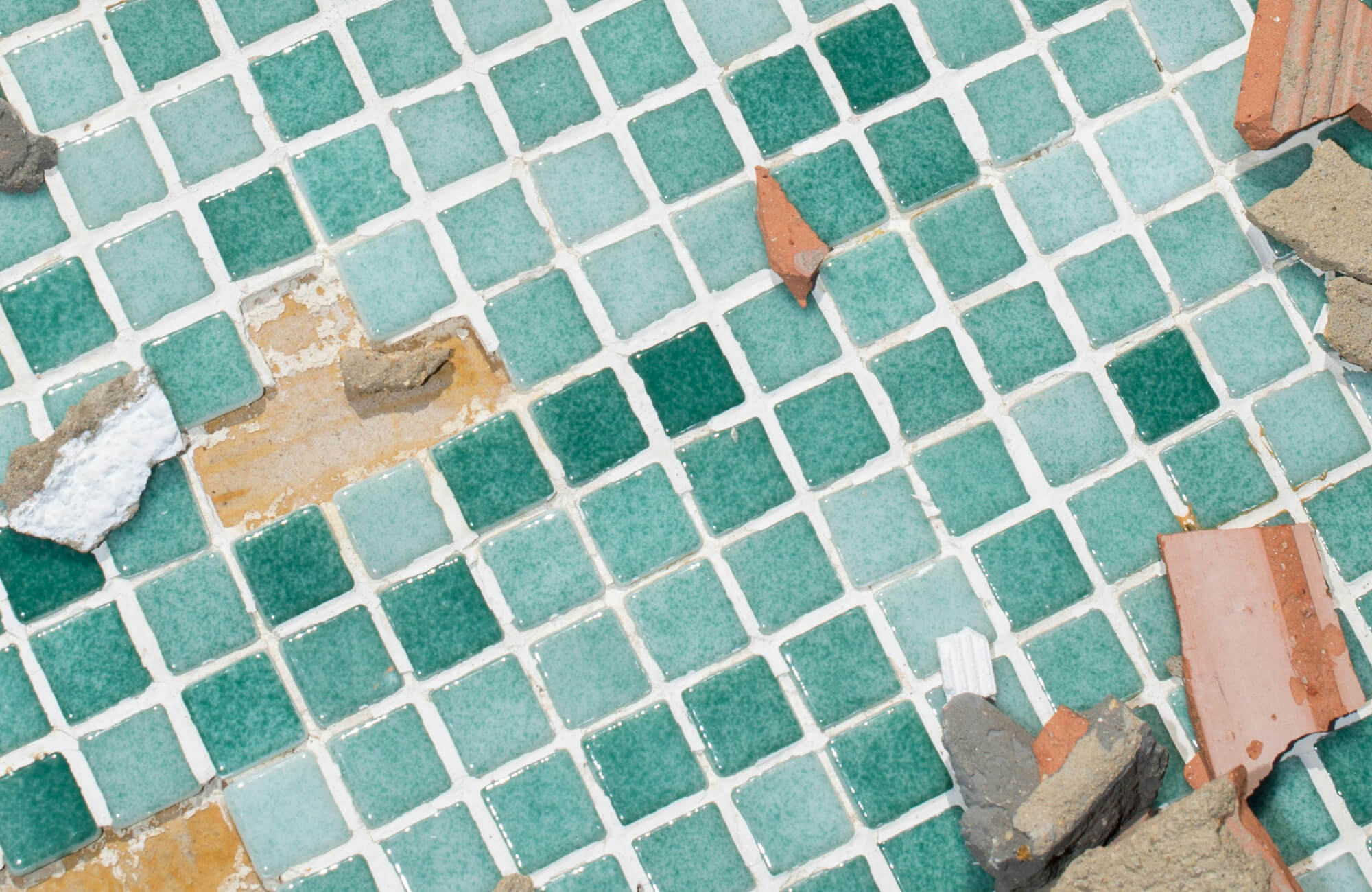 Damaged ceramic mosaic tile floor with varying teal squares, scattered debris, and visible grout wear, and missing tiles.