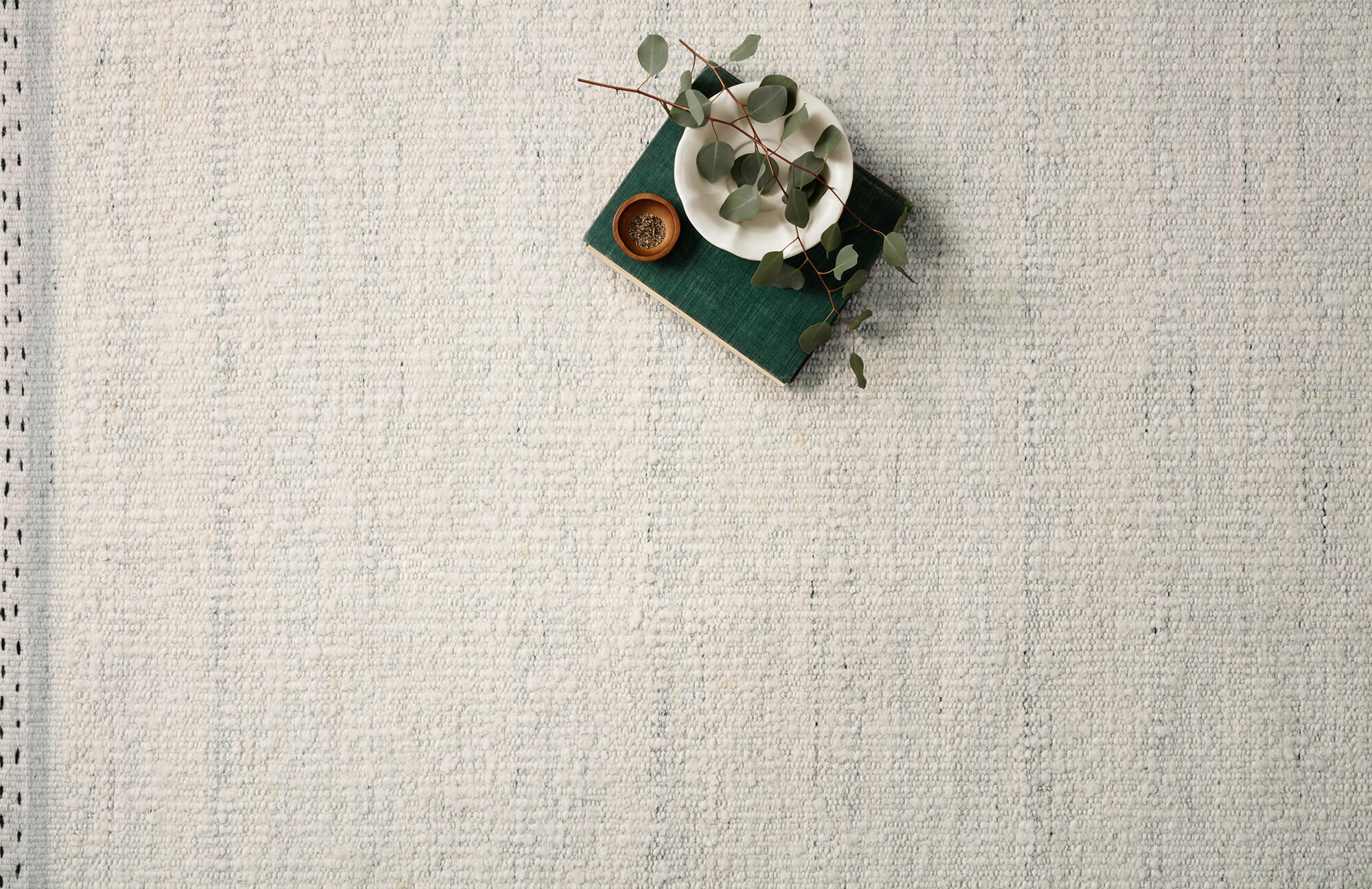 Soft ivory-toned textured rug with subtle woven detailing, styled with eucalyptus, ceramic, and a green book.