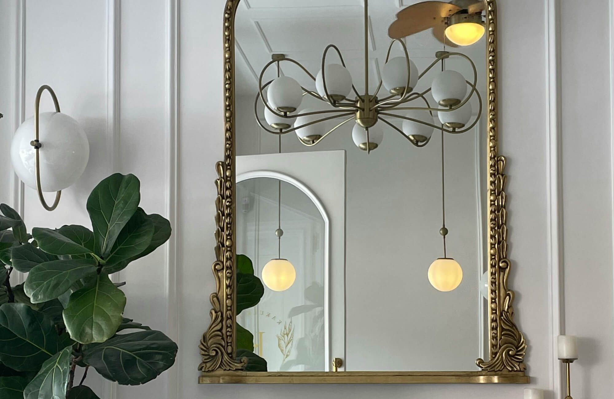 A large wall mirror with an ornate antique brass frame reflects sculptural lighting and soft architectural details, adding vintage elegance and depth to the refined interior setting.
