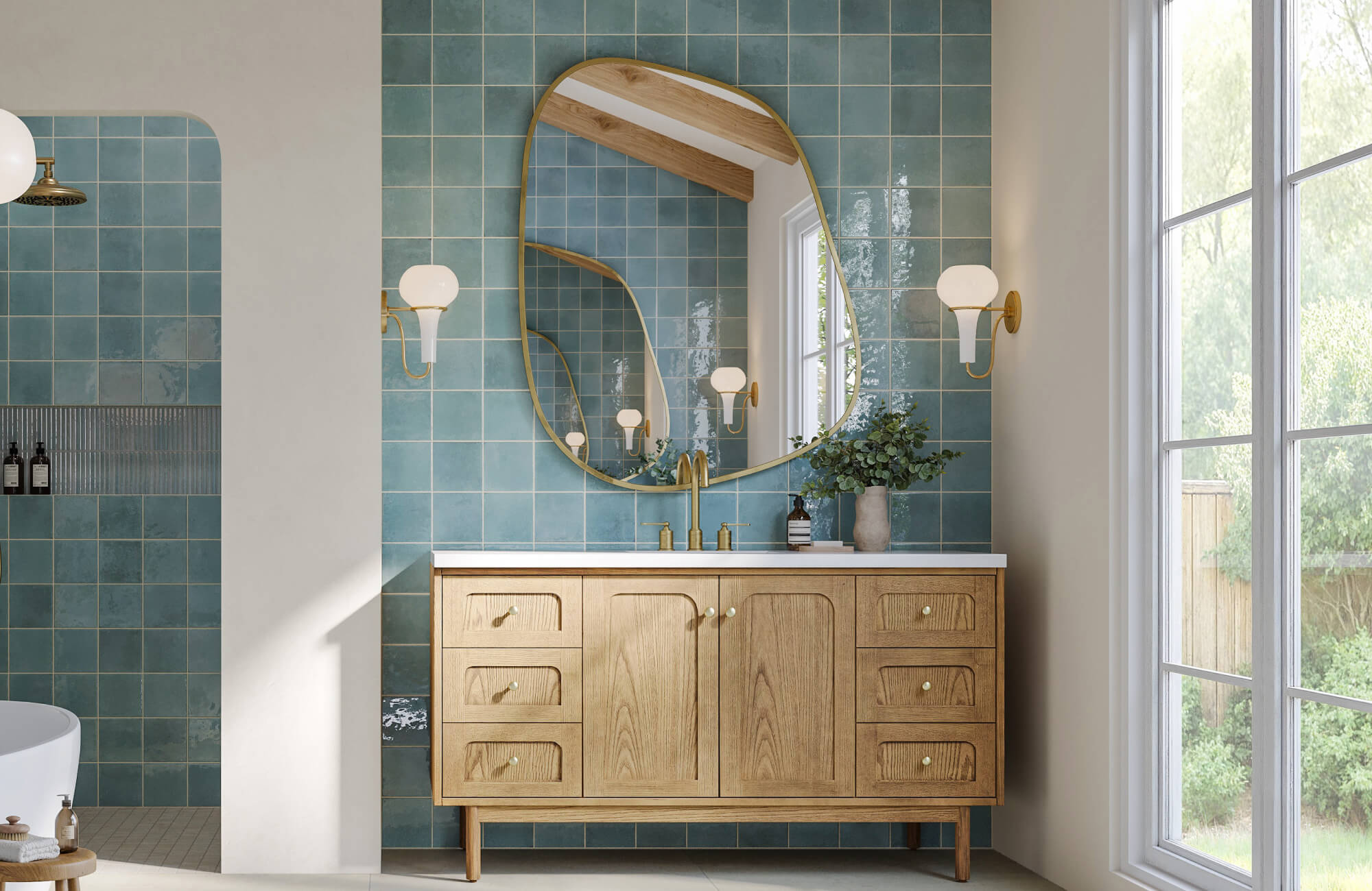 A large wall mirror with an organic rounded shape and slim brass frame reflects natural light against blue tiled walls, enhancing the bathroom’s warm wood vanity and airy, modern design.