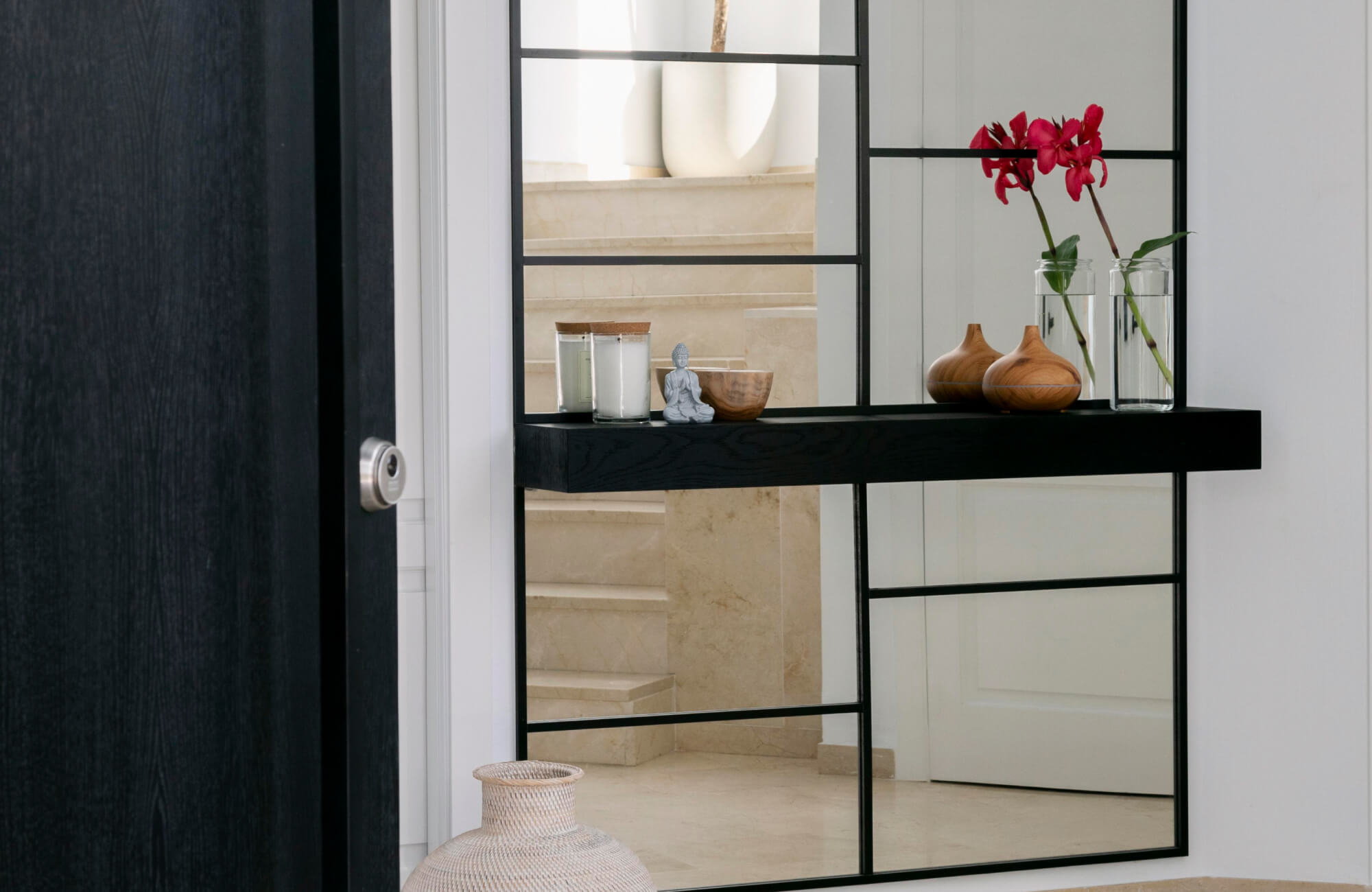 A large wall mirror with a black metal grid frame creates a striking focal point, reflecting clean architectural lines while complementing the minimalist décor and curated accessories on the floating shelf.