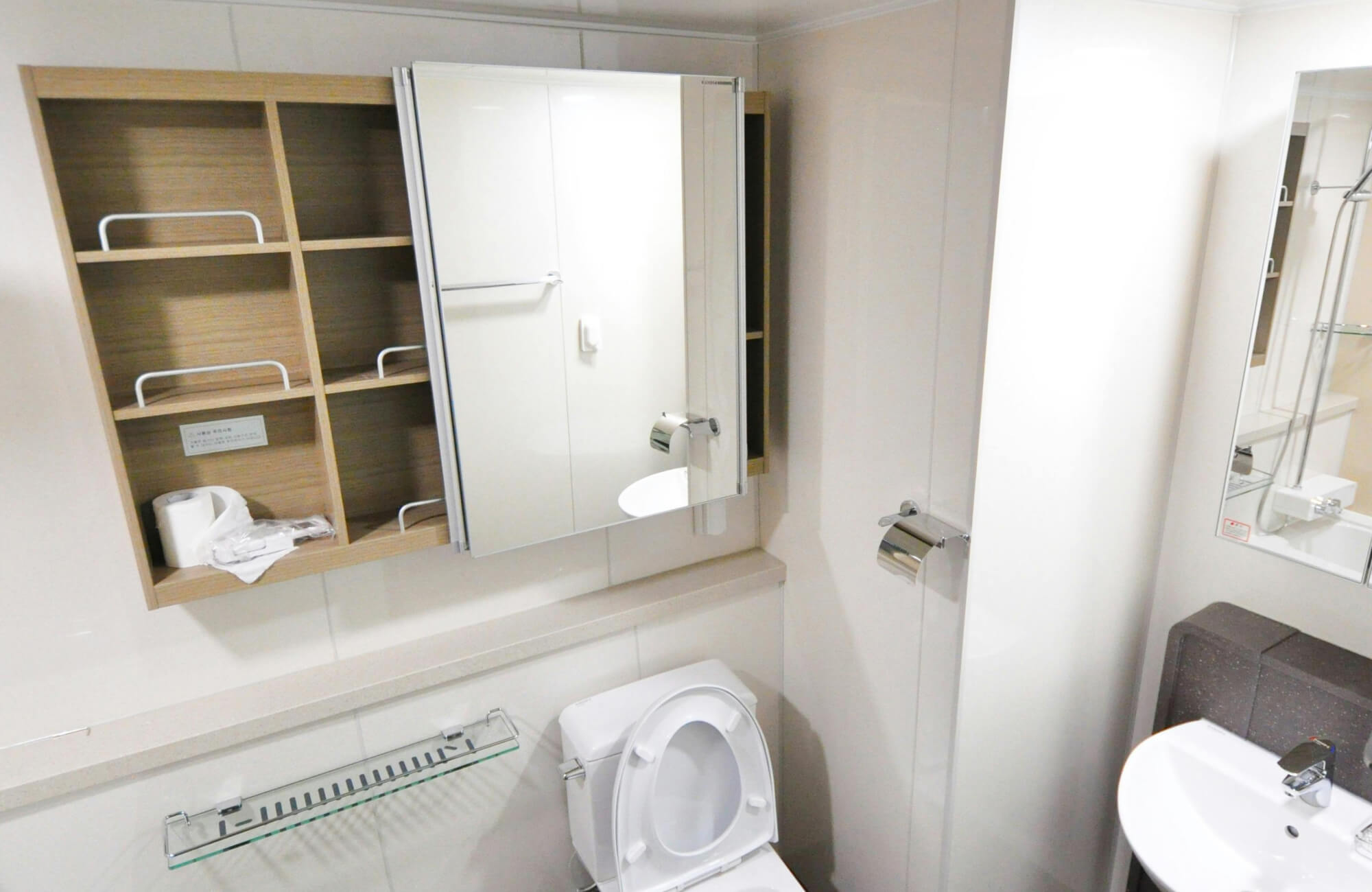 Sliding mirrored cabinet with a mix of open wood shelving and glass-front doors, installed in a compact bathroom.