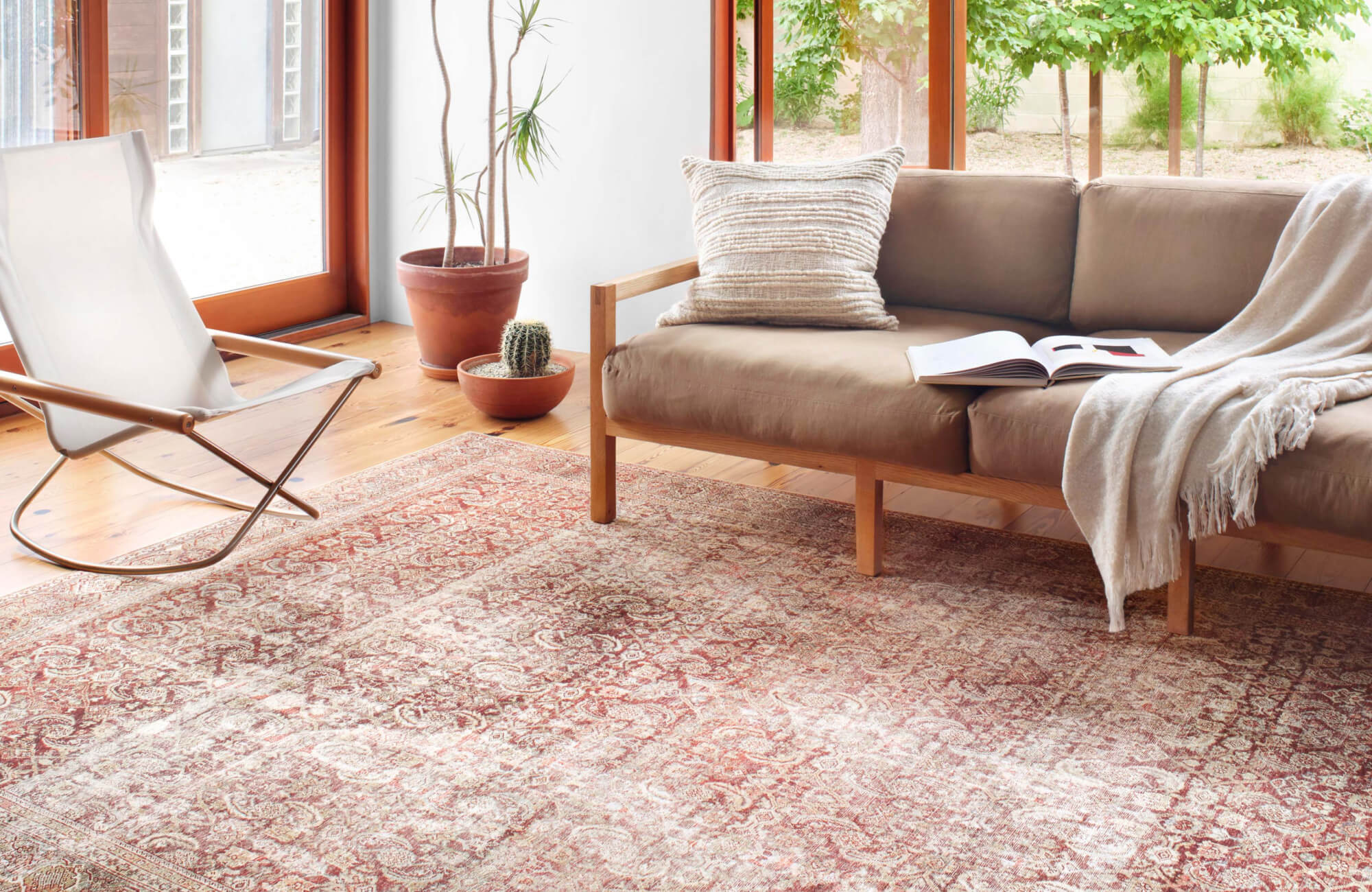 Modern living room with large red and beige patterned rug on light wood floors, tan couch, natural light, and terracotta plant pots