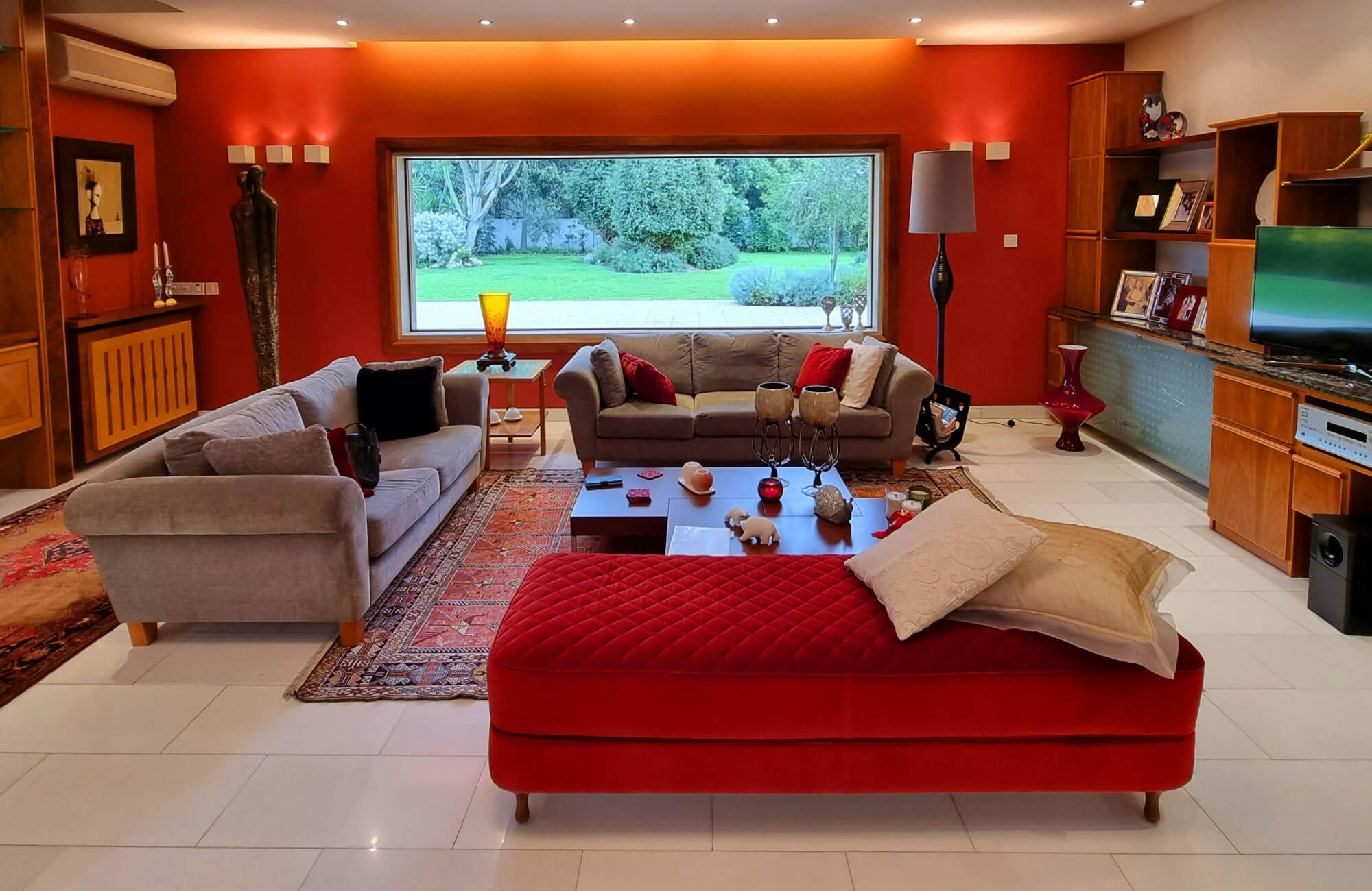 Eclectic living room with bold orange walls, layered red and orange patterned rugs, gray sofas, and red tufted bench on white tile floor