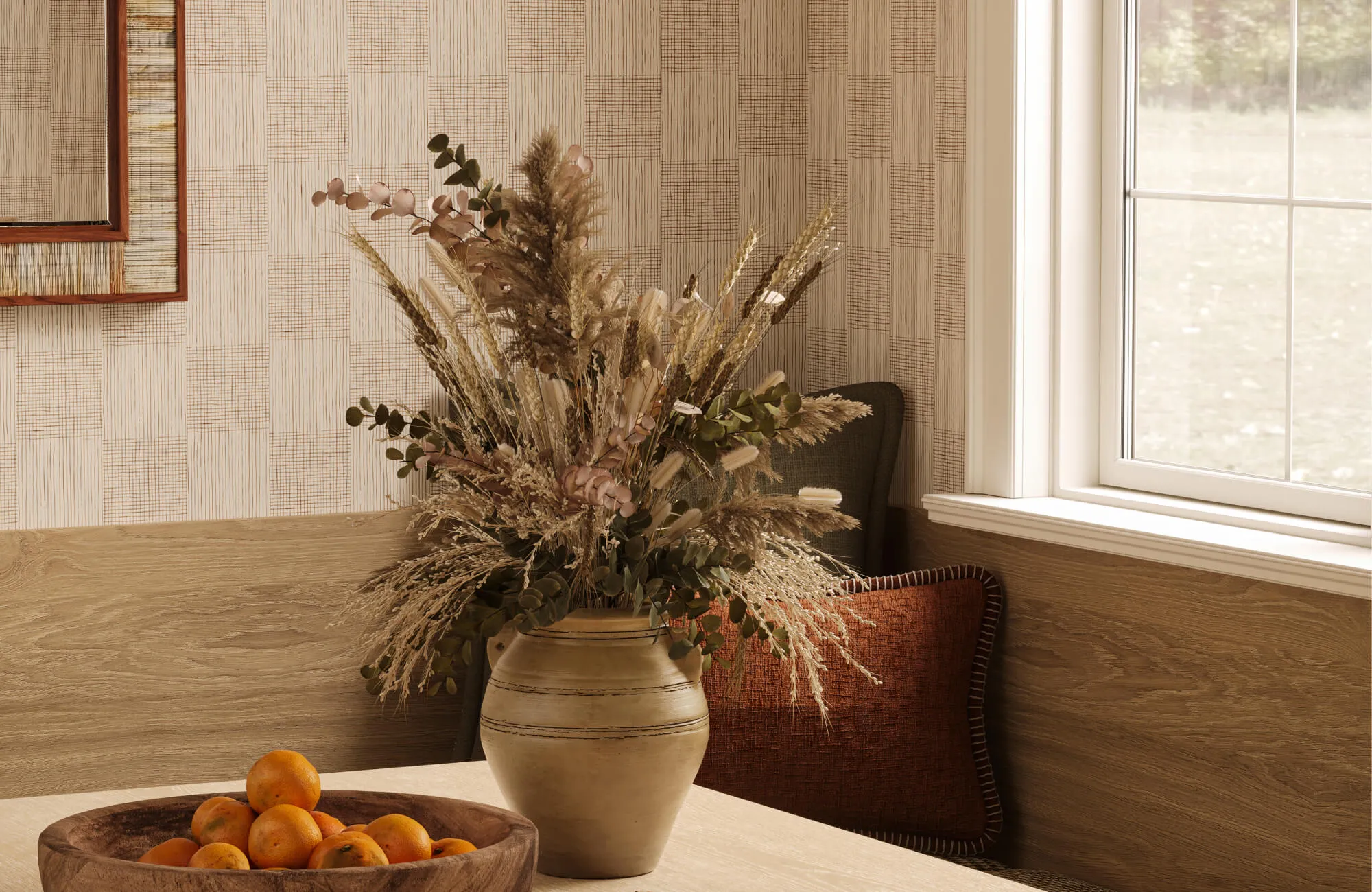 Neutral dining nook with a cream ceramic vase filled with dried grasses and greenery on a wood table beside a bowl of oranges, patterned wall covering, and warm natural light.
