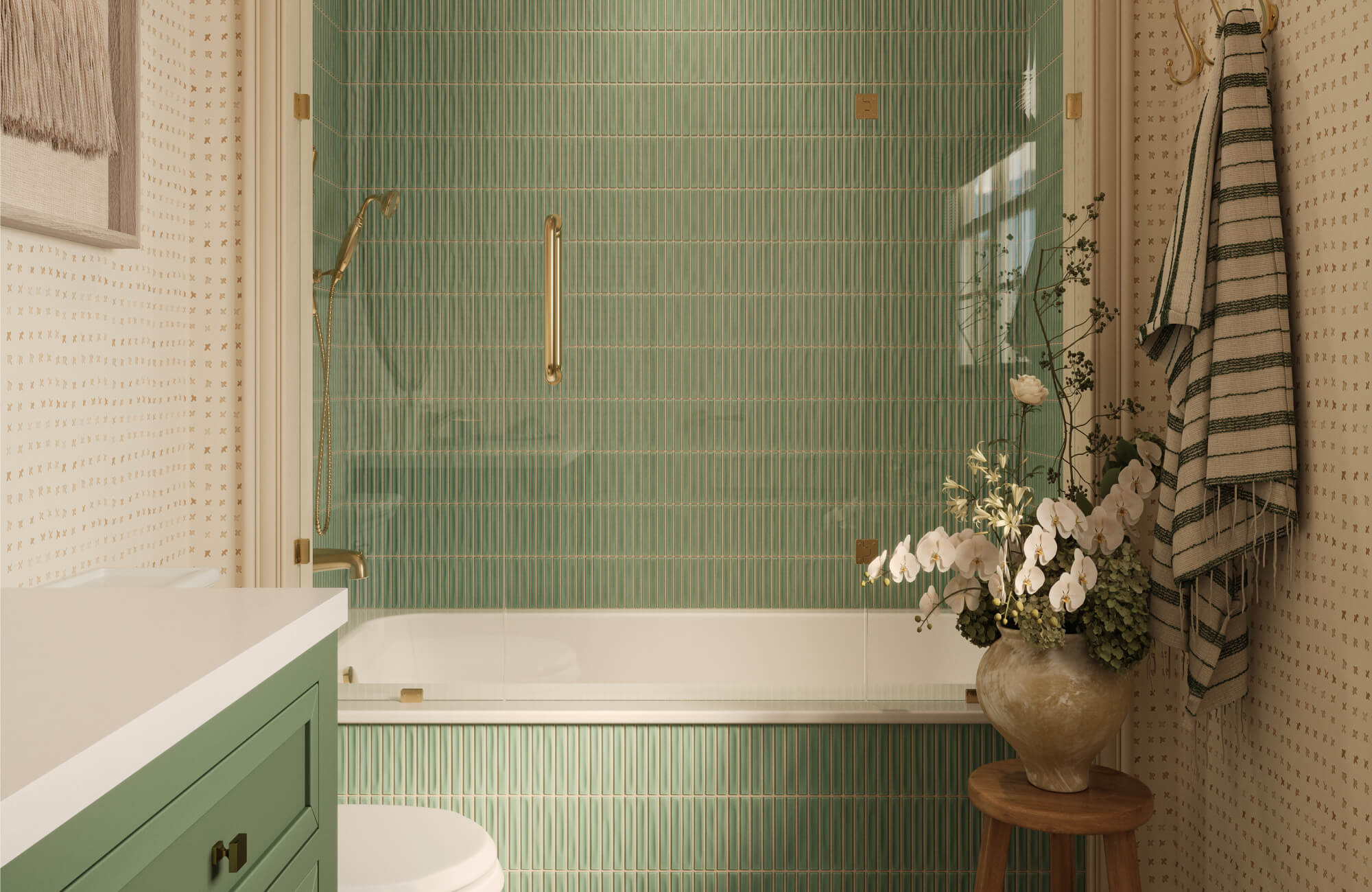 Bathroom with sage green vertical shower tile, tan patterned wallpaper, brass fixtures, striped textiles, and a large neutral vase with white flowers on a wood stool.