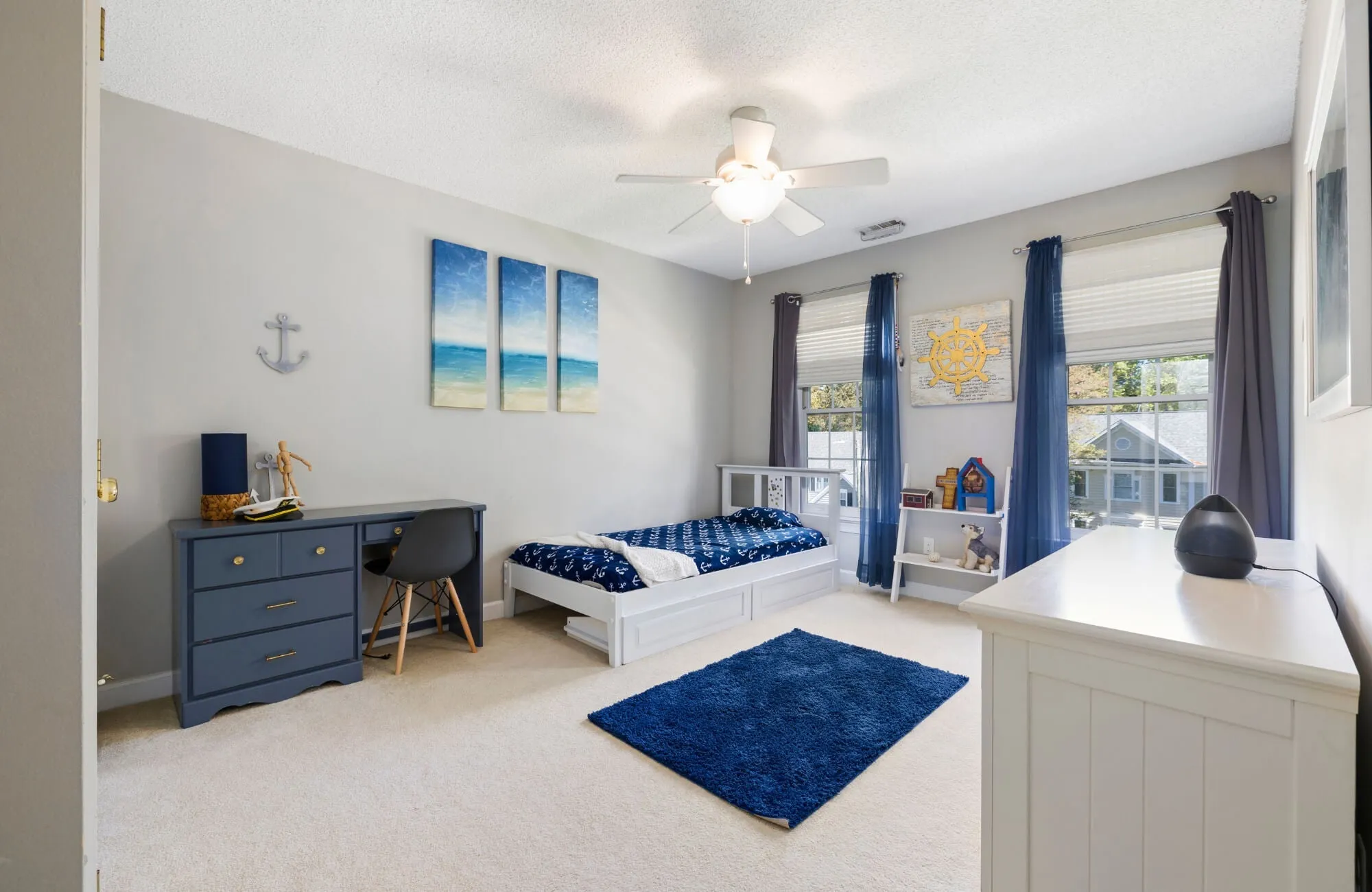 Nautical-themed bedroom with a navy blue rug, white bed, blue desk, and ocean artwork creating a bright, coastal-inspired space.