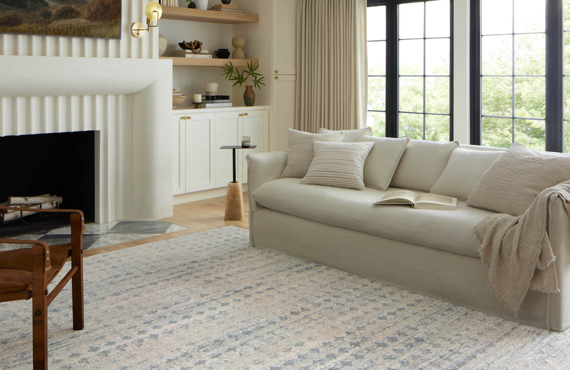 Elegant living room with a patterned blue rug, cream sofa with neutral pillows, modern fireplace, and large windows bringing in natural light.