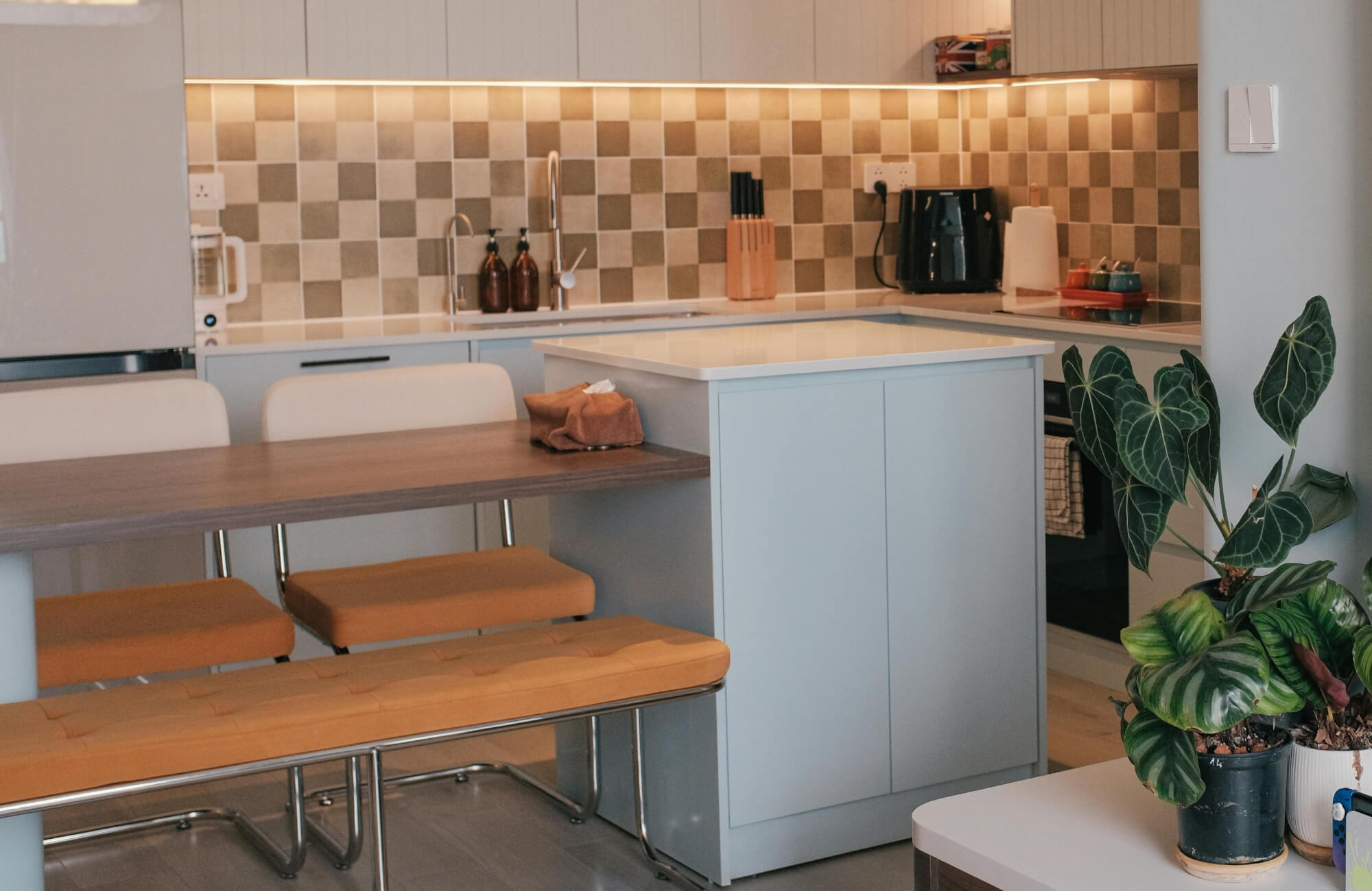 Modern kitchen with neutral checkerboard backsplash tile and sleek white countertops paired with light blue cabinetry and warm-toned seating