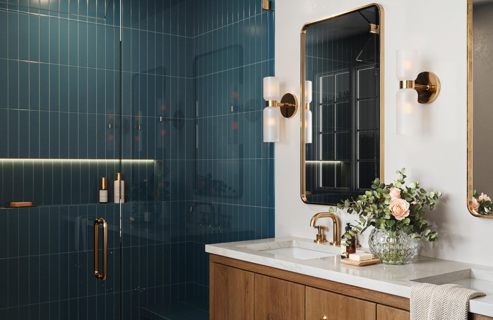 null Bathroom featuring rich teal blue ceramic tile in vertical stack pattern, brass fixtures, modern sconces, and dual vanity with floral décor