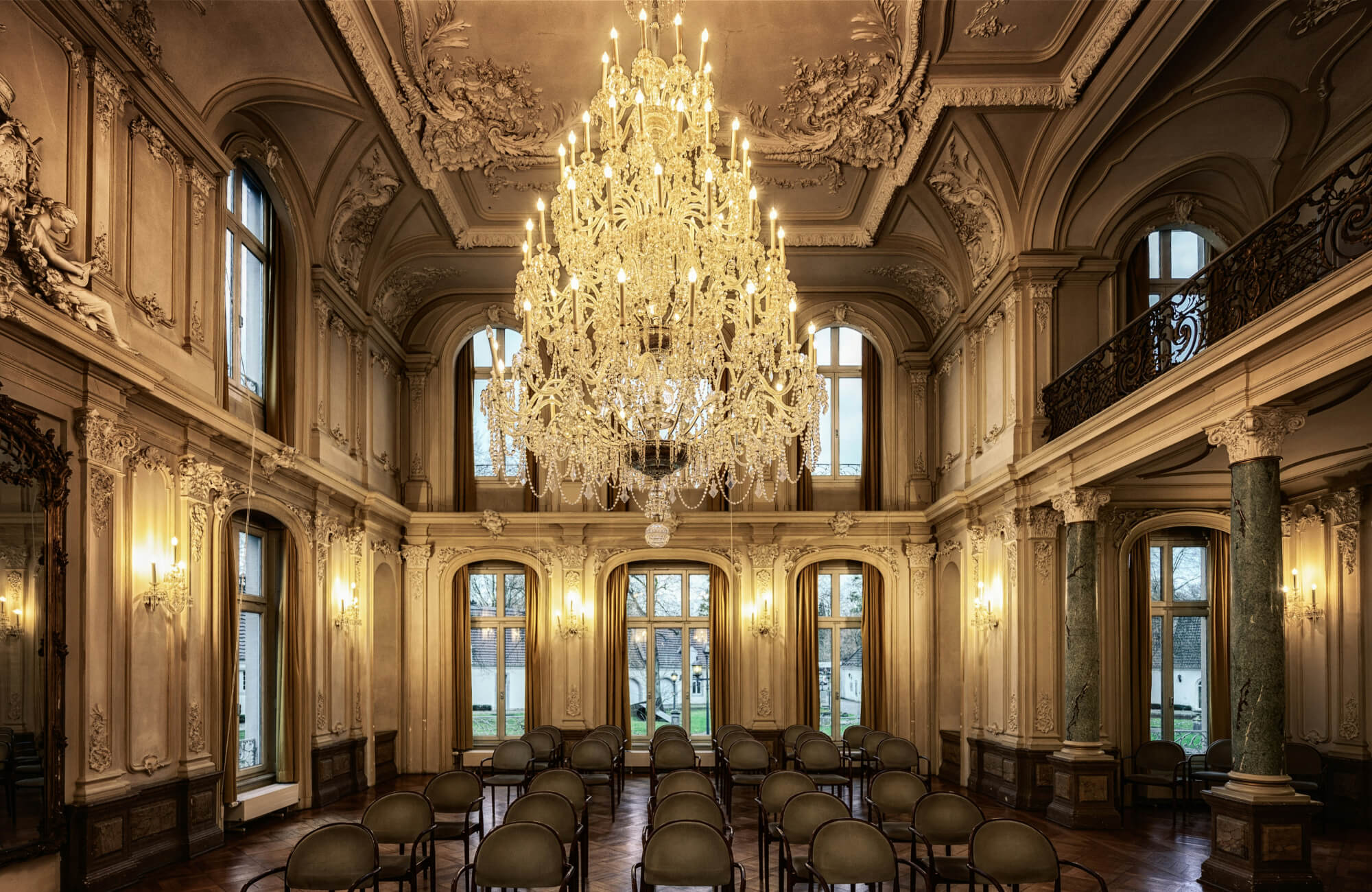 null Grand ballroom with an exquisite crystal chandelier illuminating the ornate architecture, elegant wall moldings, and rows of classic seating beneath towering windows.