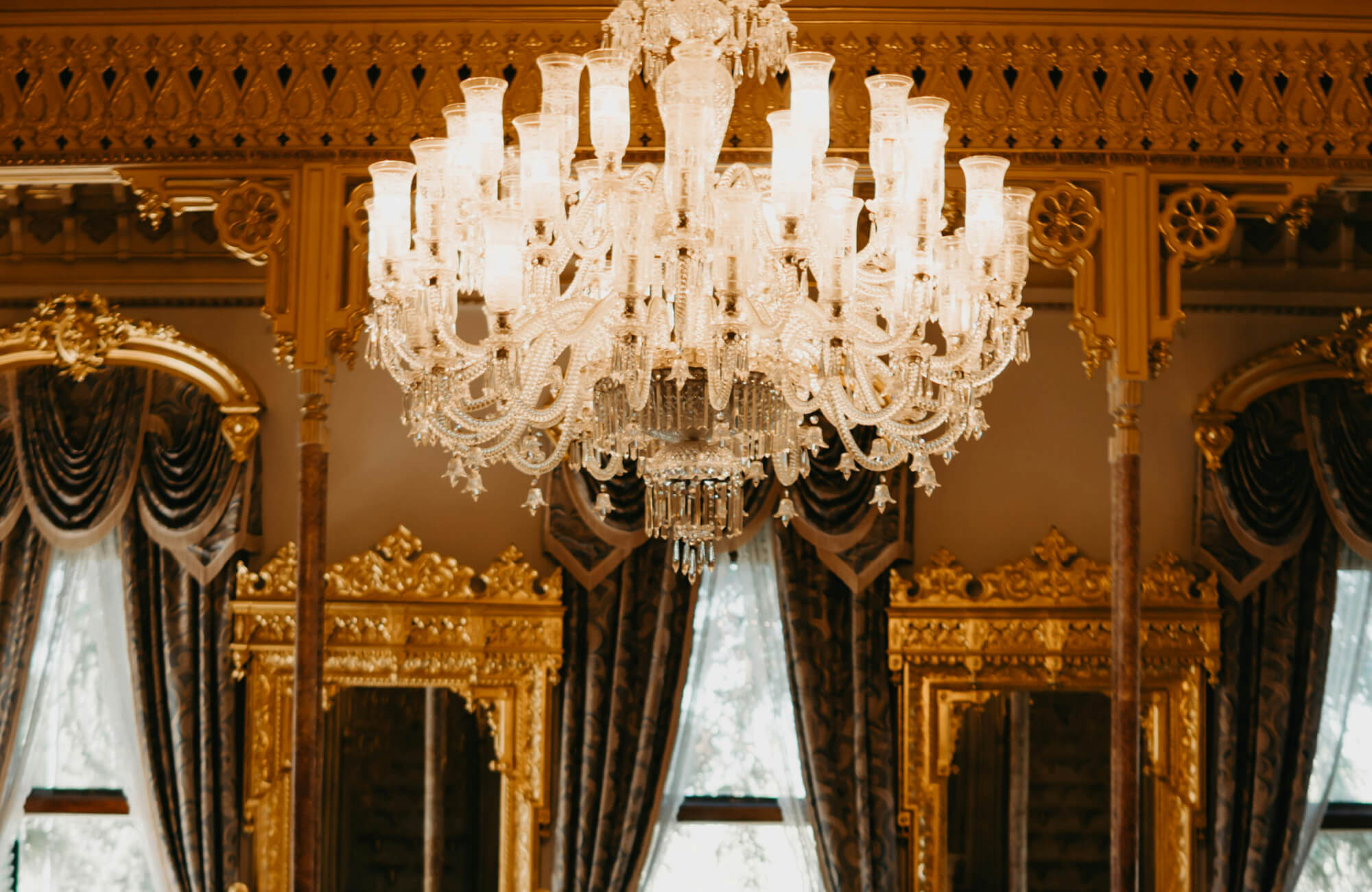 null Elegant crystal chandelier with intricate glass arms and glowing shades, enhancing the lavish golden décor and regal drapery of the grand interior.