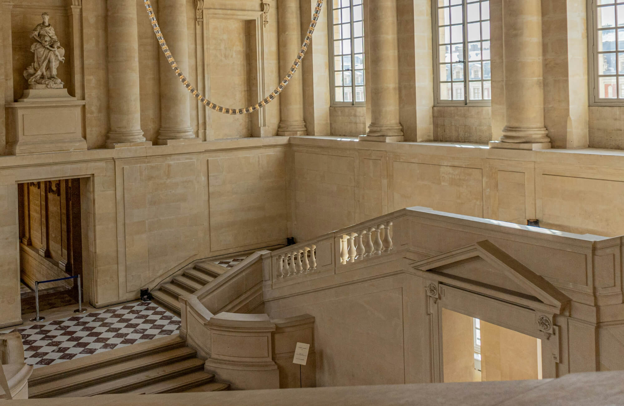 null Grand staircase inside a historic building with beige stone walls, large windows, and a checkerboard tile floor at the entrance.