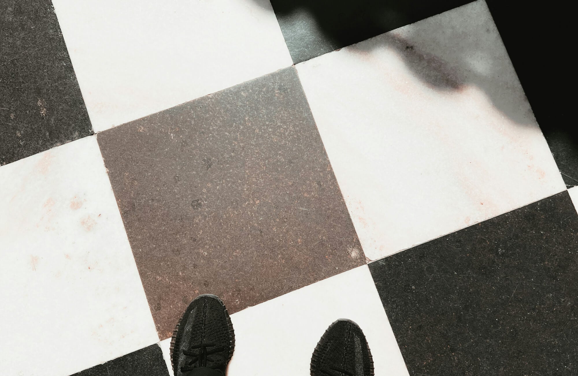 null Checkerboard floor tiles in black and white with a person's black shoes partially visible, casting a shadow.