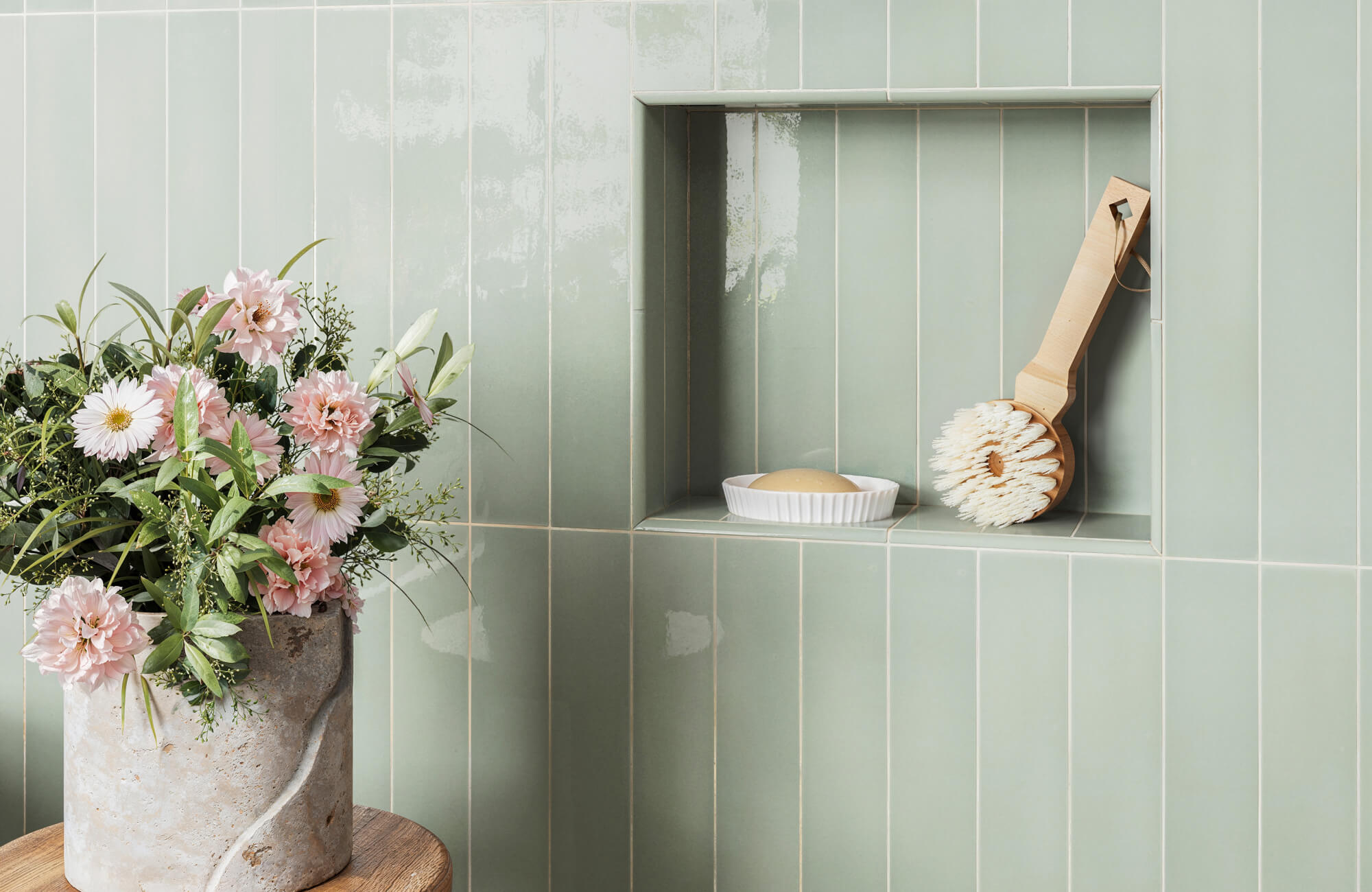Vertically stacked soft pistachio green subway tiles with a built-in niche holding a wooden bath brush and soap, complemented by a vase of pink flowers.