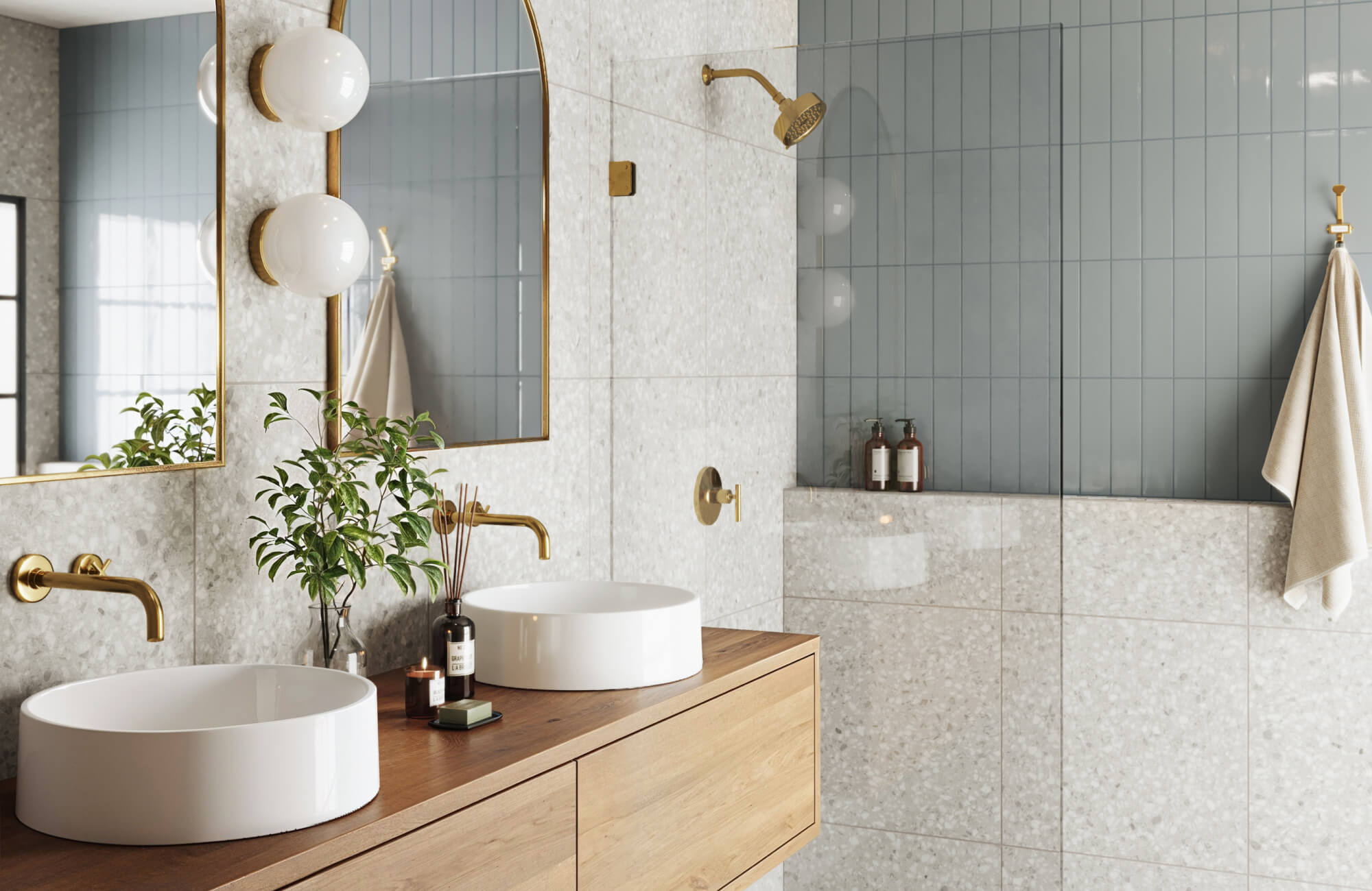 Sleek muted blue-green subway tiles pair with terrazzo look walls, a wooden vanity, gold fixtures, and twin round sinks for a modern bathroom design.