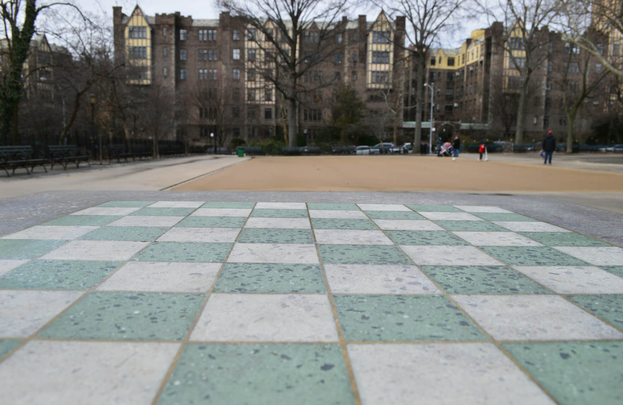 Outdoor stone checkerboard with green and white floor tiles grouted in orange-brown in a city park, with classic buildings and bare trees in the background.