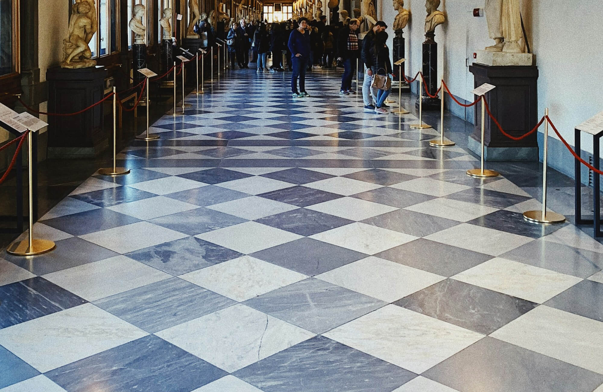 Historic gallery hallway featuring large blue-gray and white floor tiles laid in a diagonal checkerboard pattern, finished with bluish-gray grout lines that soften the contrast, flanked by classical sculptures and filled with visitors moving through the space.