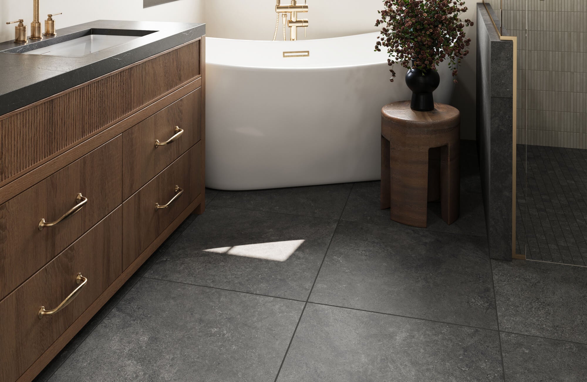 null Contemporary bathroom with dark floor tiles, a white tub, wood vanity, and a floral arrangement on a small wooden stool.