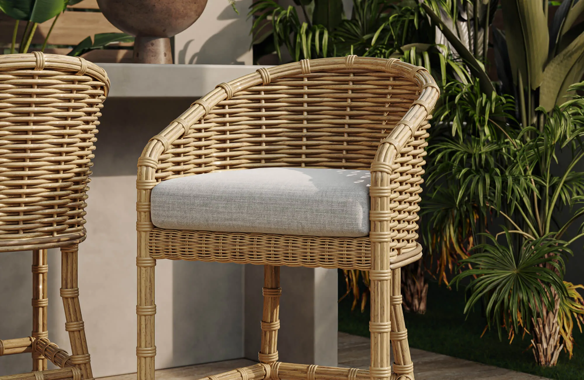 Natural woven outdoor counter stool with a light gray cushion beside a patio counter, surrounded by lush green plants and warm sunlight.