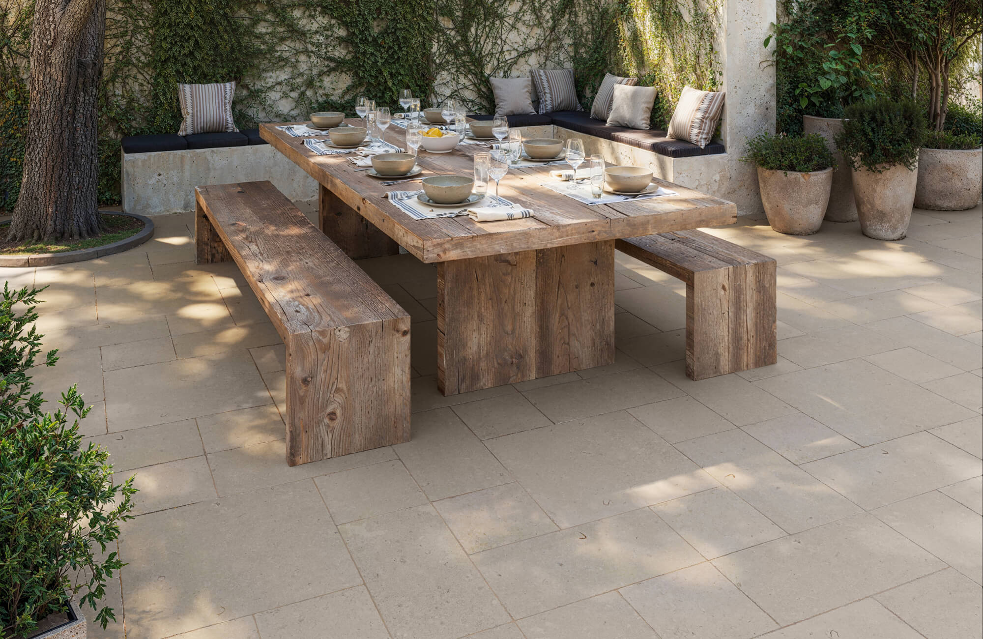 A rustic outdoor dining area showcases warm beige outdoor tile flooring arranged in a modular pattern beneath a large wooden table and benches, surrounded by lush greenery and built-in seating.