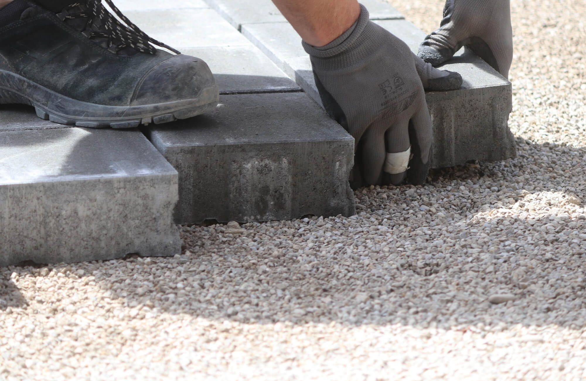 A close-up view shows a worker installing thick concrete outdoor tile pavers over a compacted gravel base, using gloved hands and protective footwear to position each piece securely.