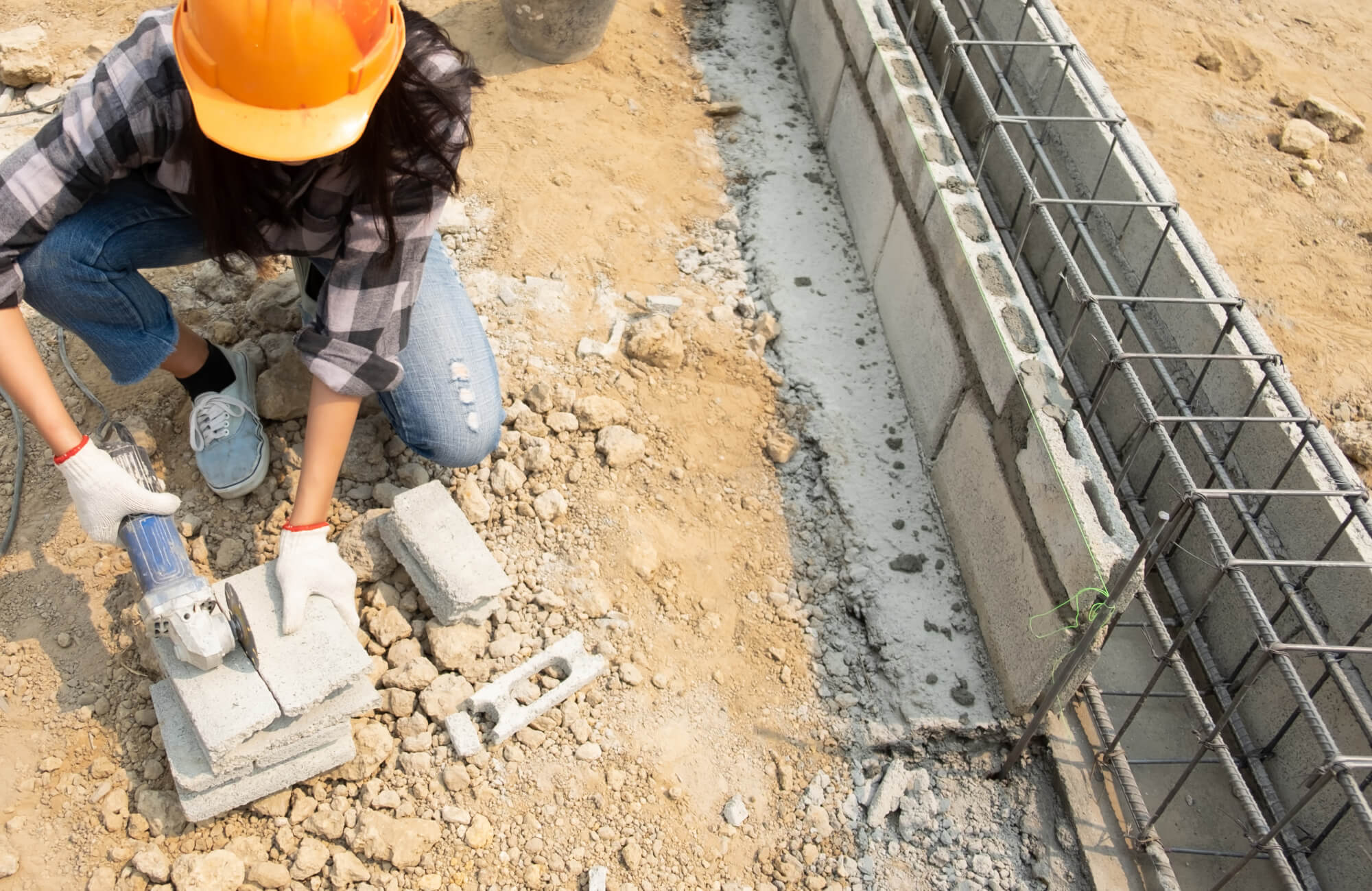 A construction worker wearing safety gear uses a power tool to cut concrete blocks beside a reinforced trench, preparing the foundation area where outdoor tile or hardscape materials may later be installed.