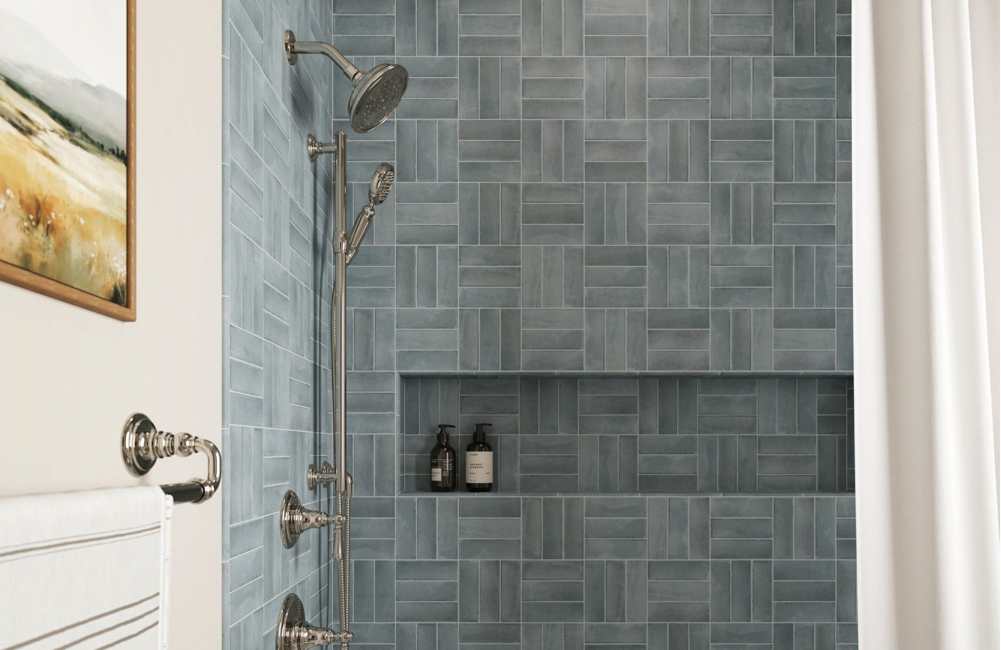 A shower wall features small blue-gray rectangular tiles arranged in a basketweave pattern with consistent light grout lines around a recessed niche and polished chrome fixtures.