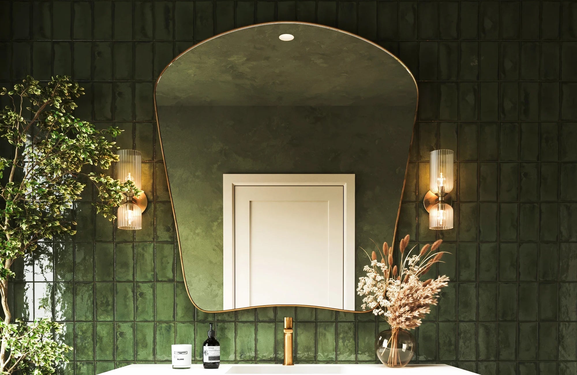 A bathroom vanity wall is covered in glossy dark green square tiles with tight grout spacing, framing a large curved mirror, warm sconces, a brass faucet, and decorative greenery.