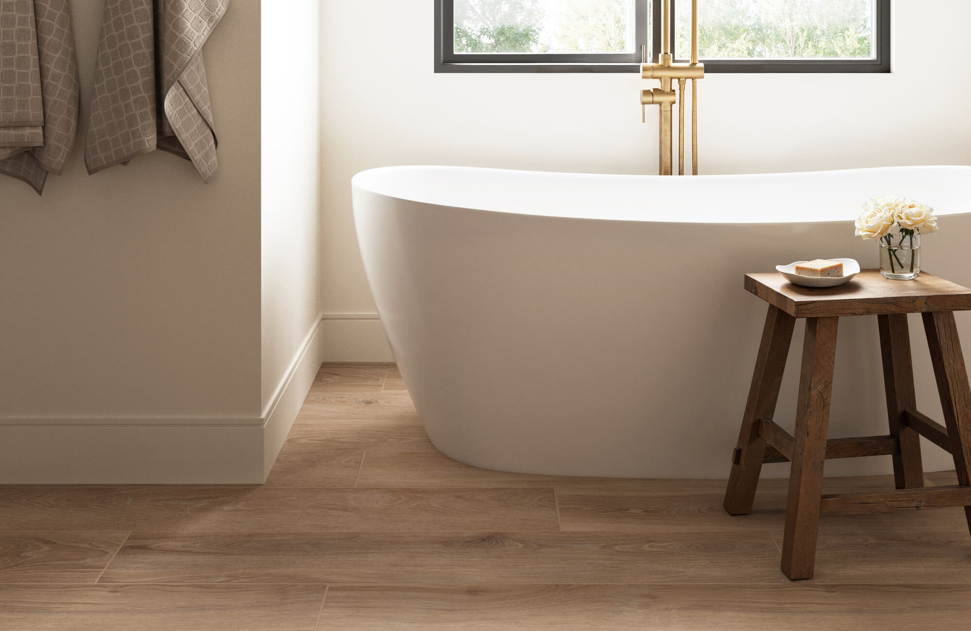 null Warm brown wood look tiles run across the bathroom floor, pairing naturally with a white freestanding tub, brass fixtures, and a small wooden stool.
