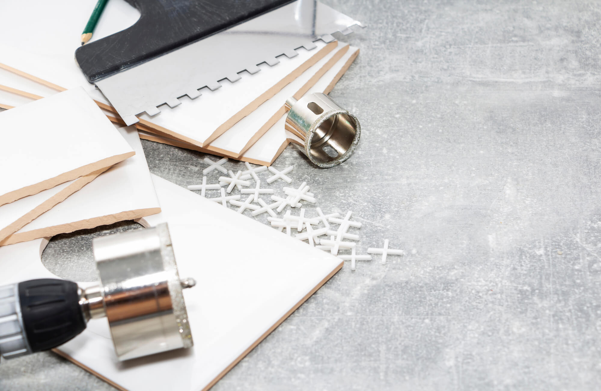 null Subway tile installation tools include a notched trowel, hole saw, spacers, and white ceramic tiles on a concrete work surface.