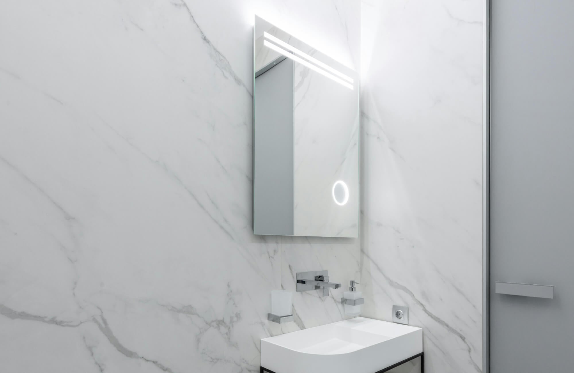 Contemporary bathroom with backlit LED smart mirror, sleek wall-mounted faucet, white rectangular sink, and marble-look wall panels