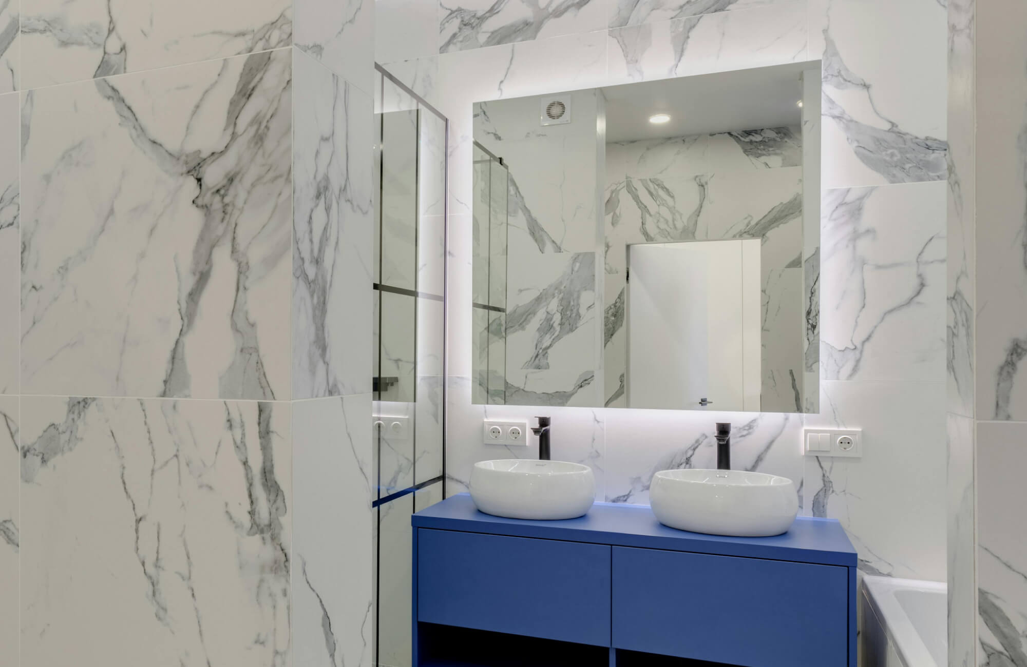 Contemporary bathroom mirror with backlit illumination reflects sleek marble walls, paired with a bold blue vanity and twin vessel sinks for a striking modern design.