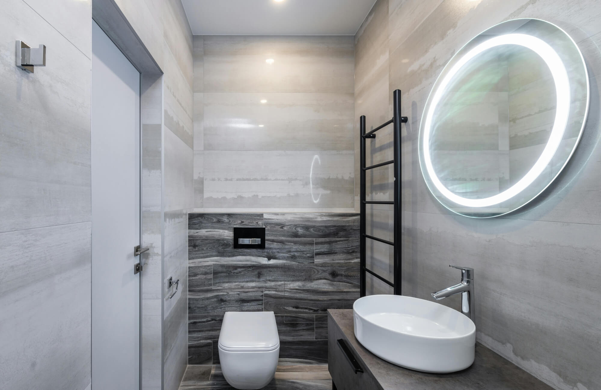 null Brightly lit round mirror illuminating a contemporary gray bathroom with sleek fixtures.