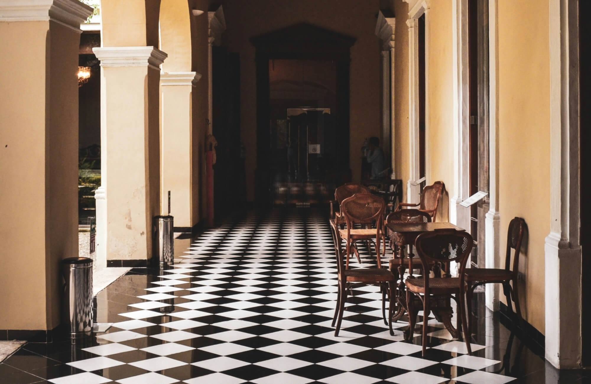 null Elegant hallway with black and white checkerboard floor tiles, lined with wooden chairs and tables, featuring high arches and cream-colored walls