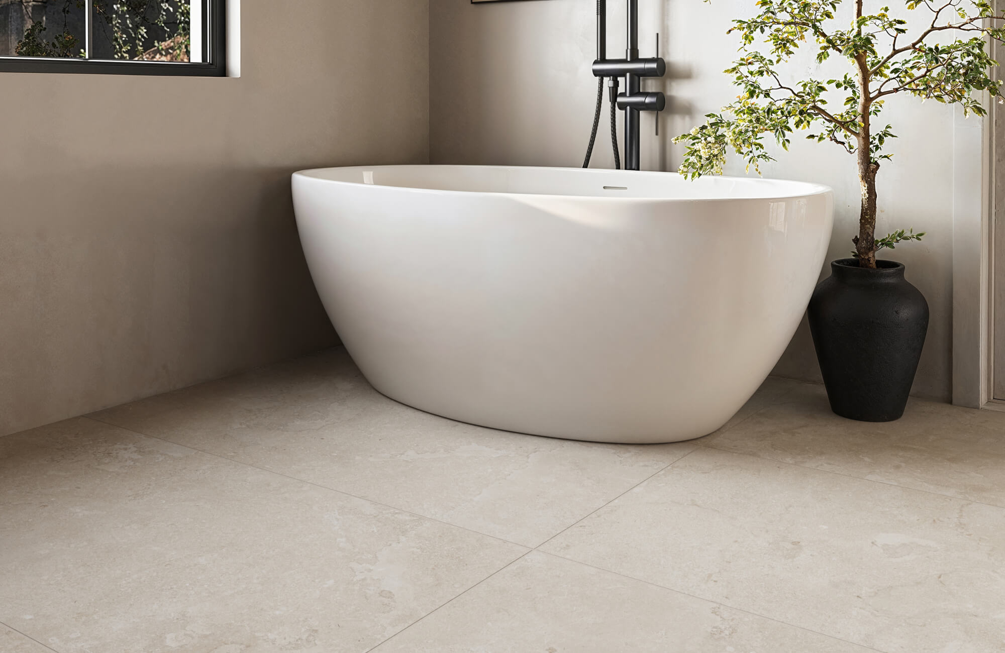 Minimalist bathroom with freestanding white tub, beige stone floor tiles, soft neutral walls, and a potted tree for a calming spa feel.