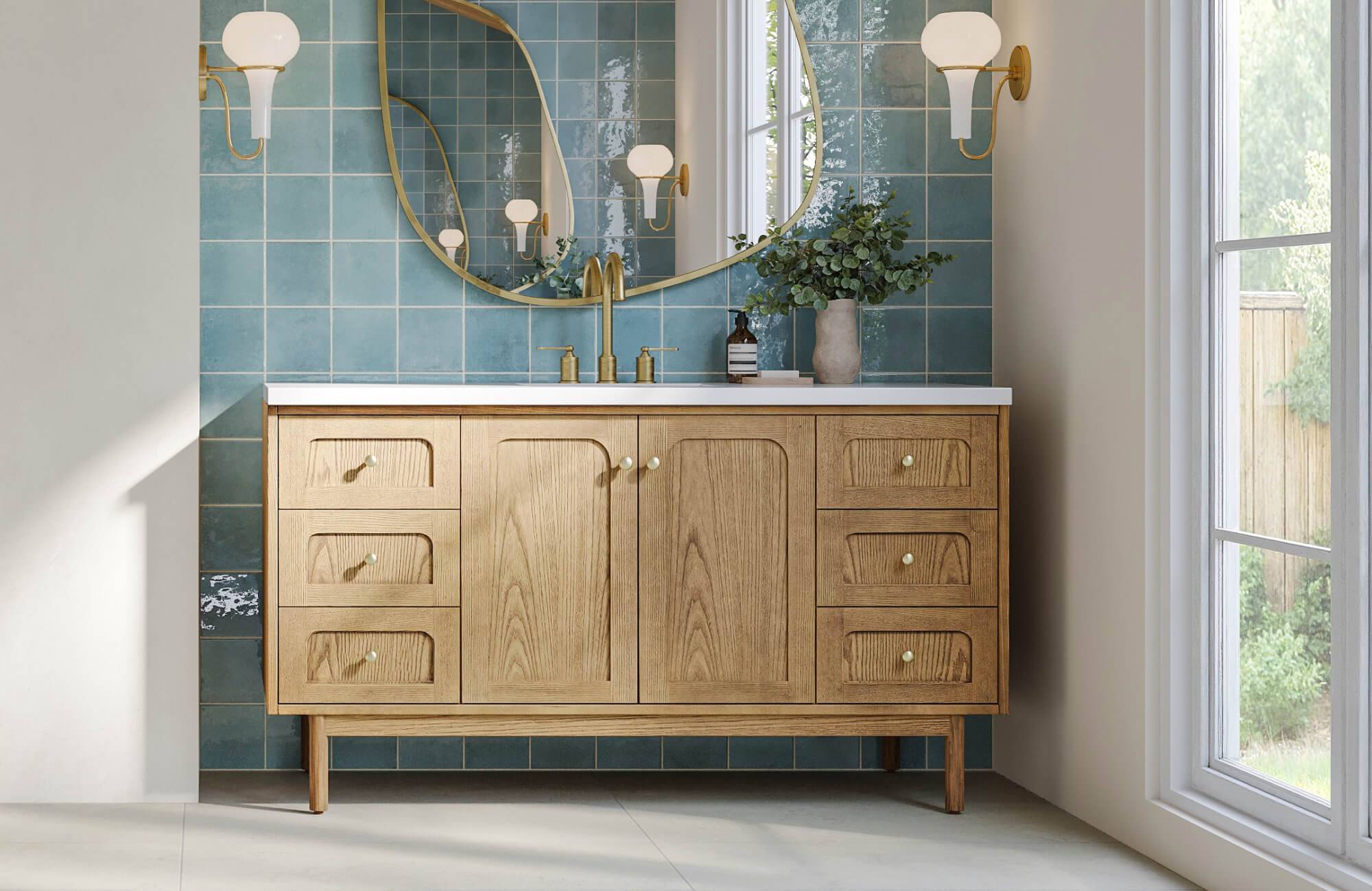 Bathroom with light oak vanity cabinet and brass fixtures set against glossy sea-green square tile wall and arched gold-framed mirror
