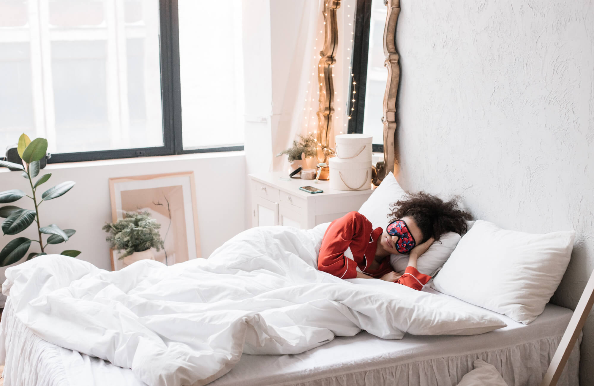 null Woman in a red pajama top and floral sleep mask resting on a white pillow in a bright white bedroom with fairy lights and green indoor plants.