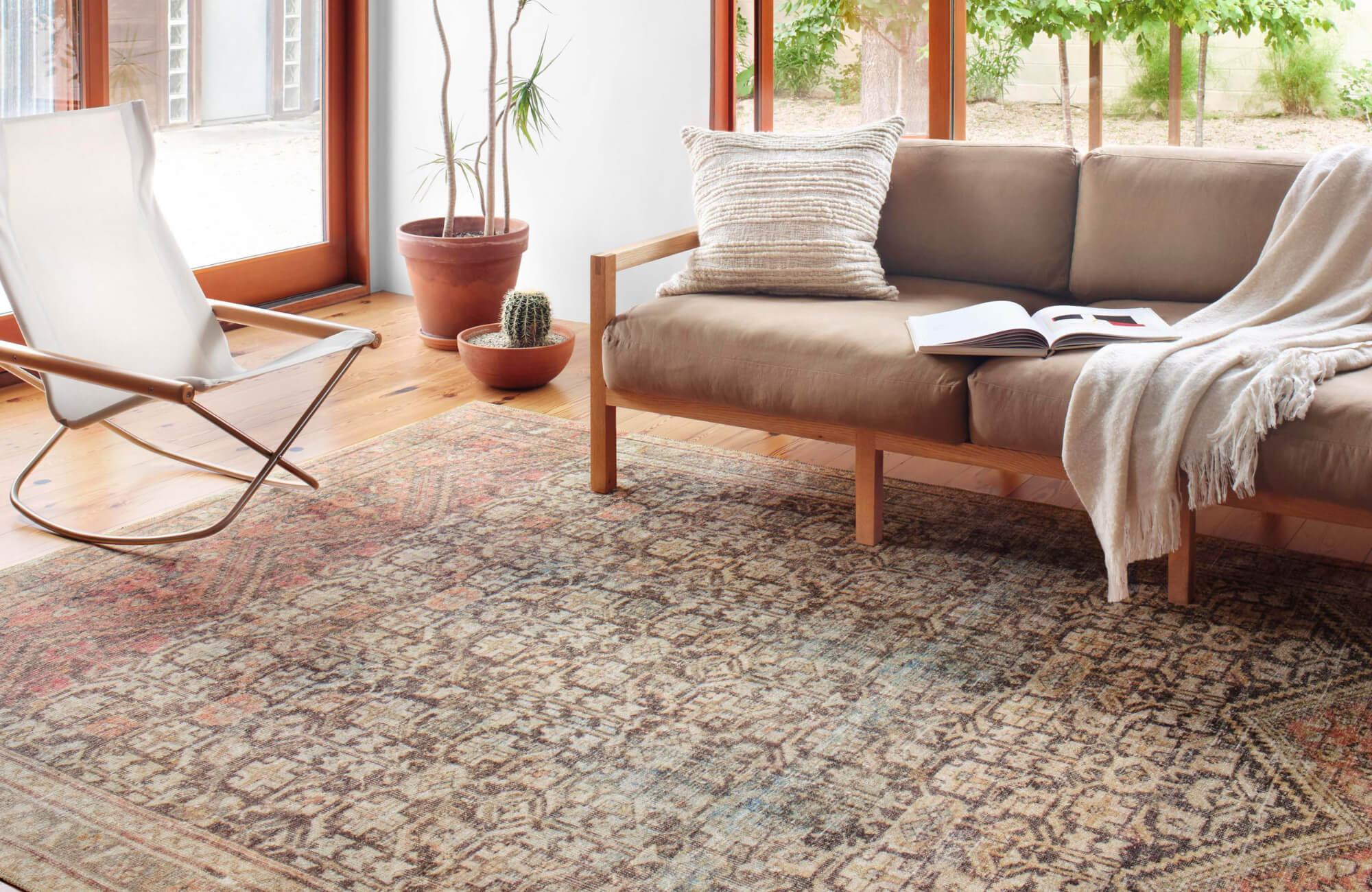 Bright living room with a tan sofa, neutral throw blanket, and patterned Moroccan rug beside large windows and potted plants.