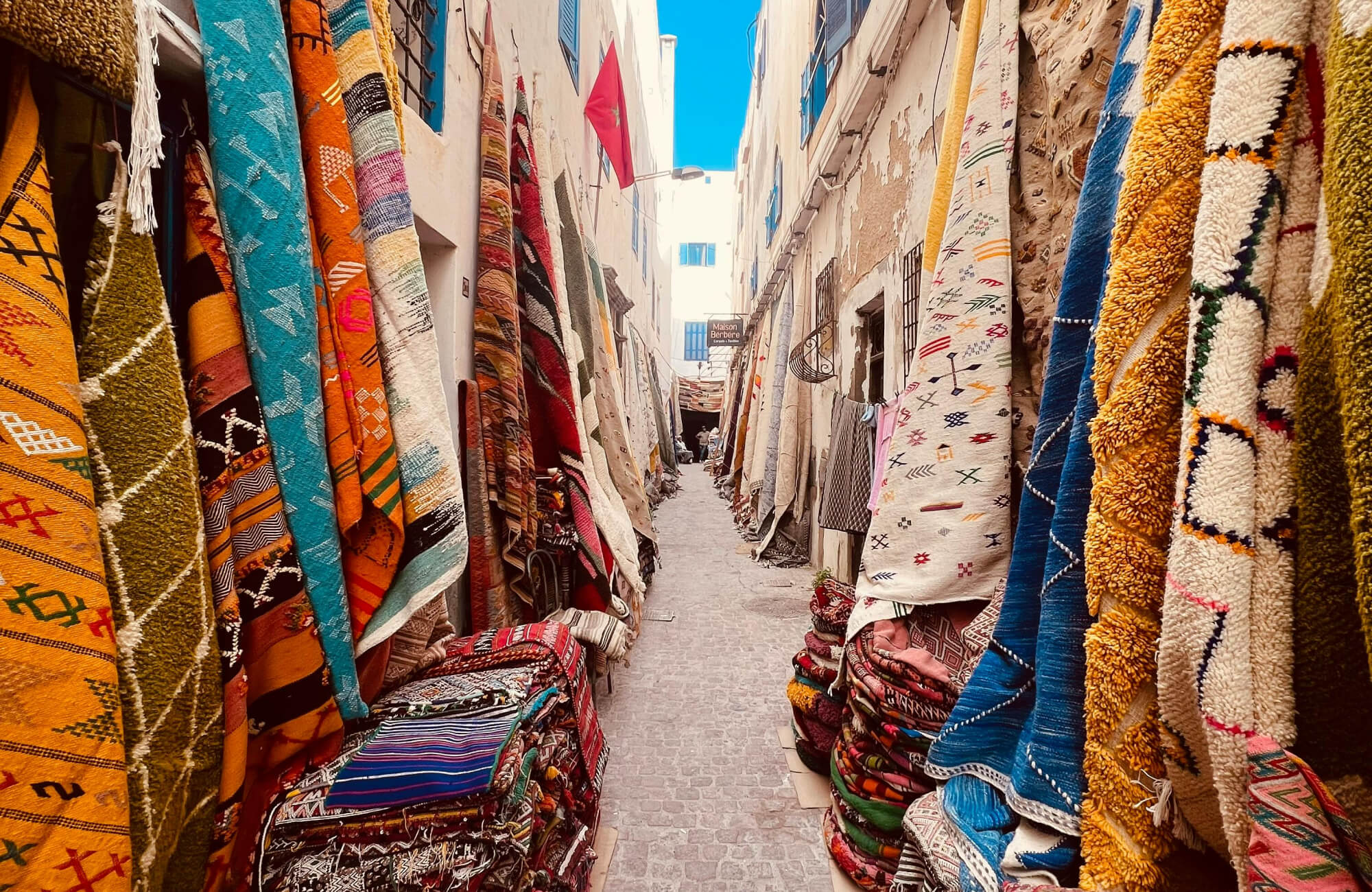 Narrow alleyway lined with vibrant Moroccan rugs in rich textures and patterns, creating a colorful, immersive marketplace scene.