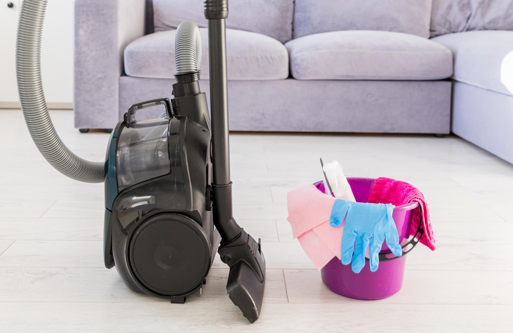 Vacuum and bucket of rug cleaning tools, including gloves, cloths, and a spray bottle, on a light floor near a sofa.