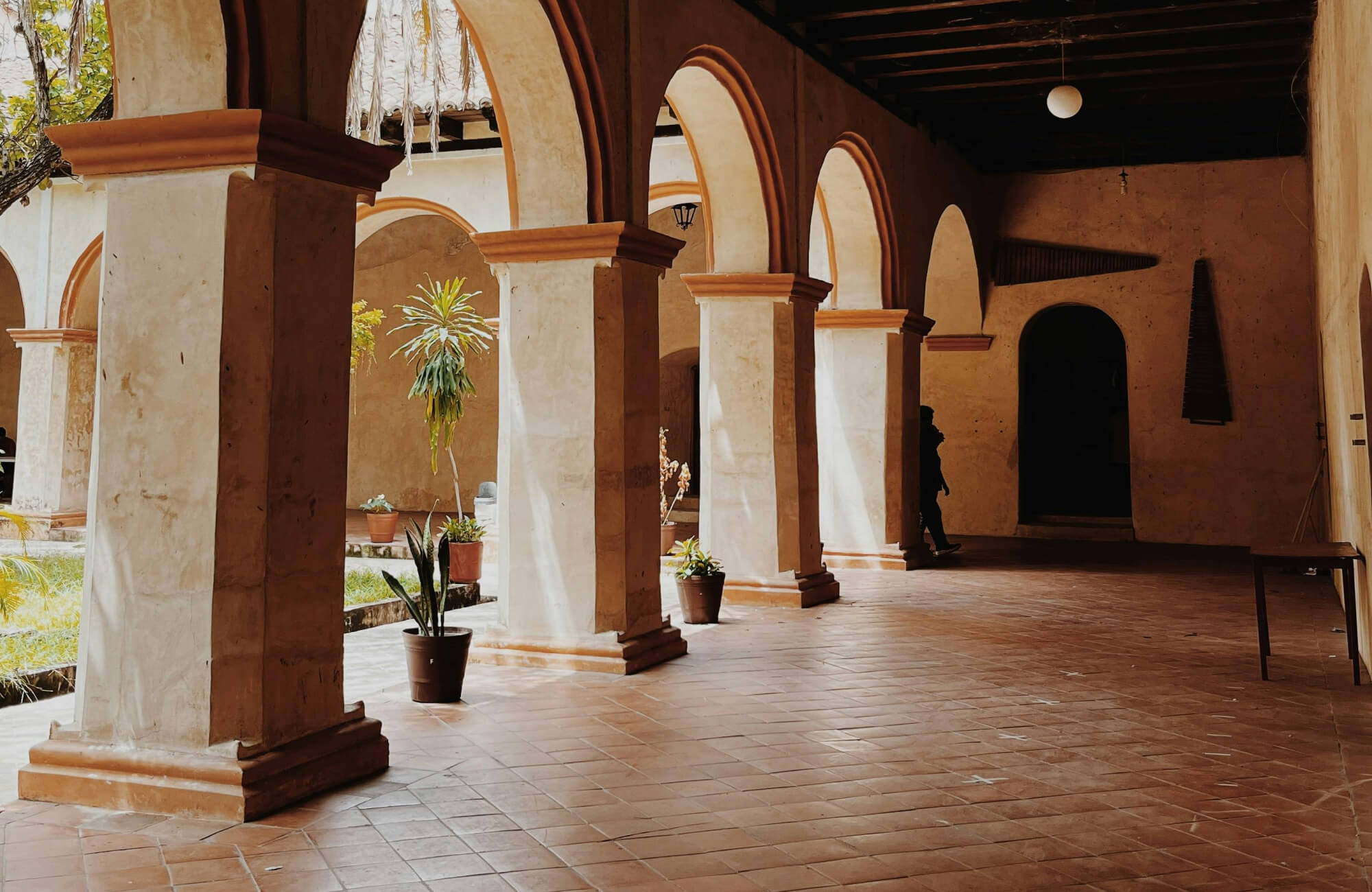 Sunlit corridor with terracotta tile flooring, arched columns, and potted plants in a historic courtyard setting.