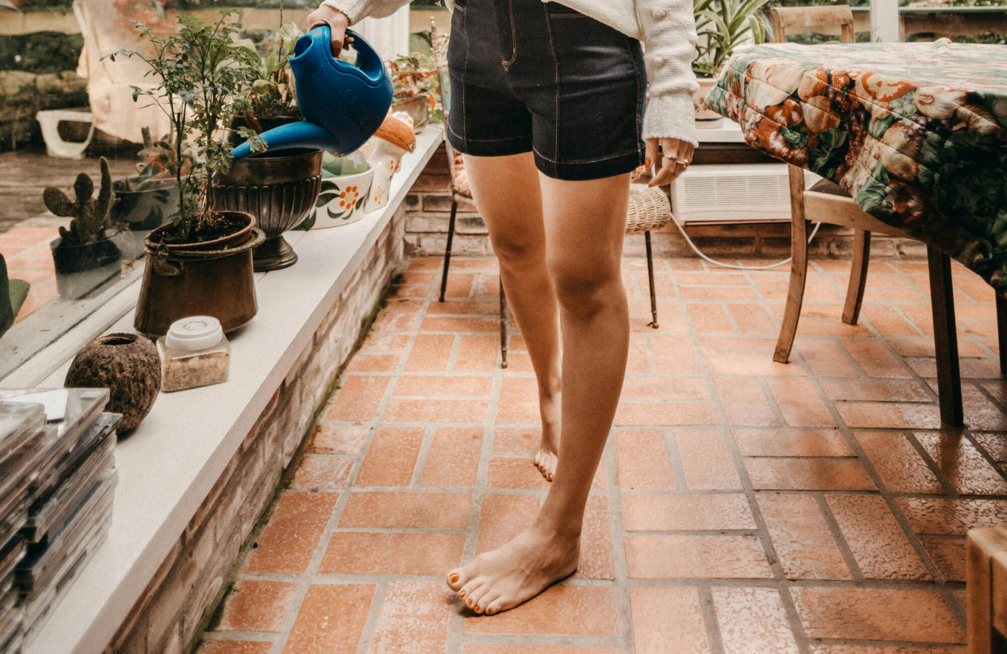 Person barefoot on red terracotta tile floor, watering potted plants near a window in a cozy indoor garden space.