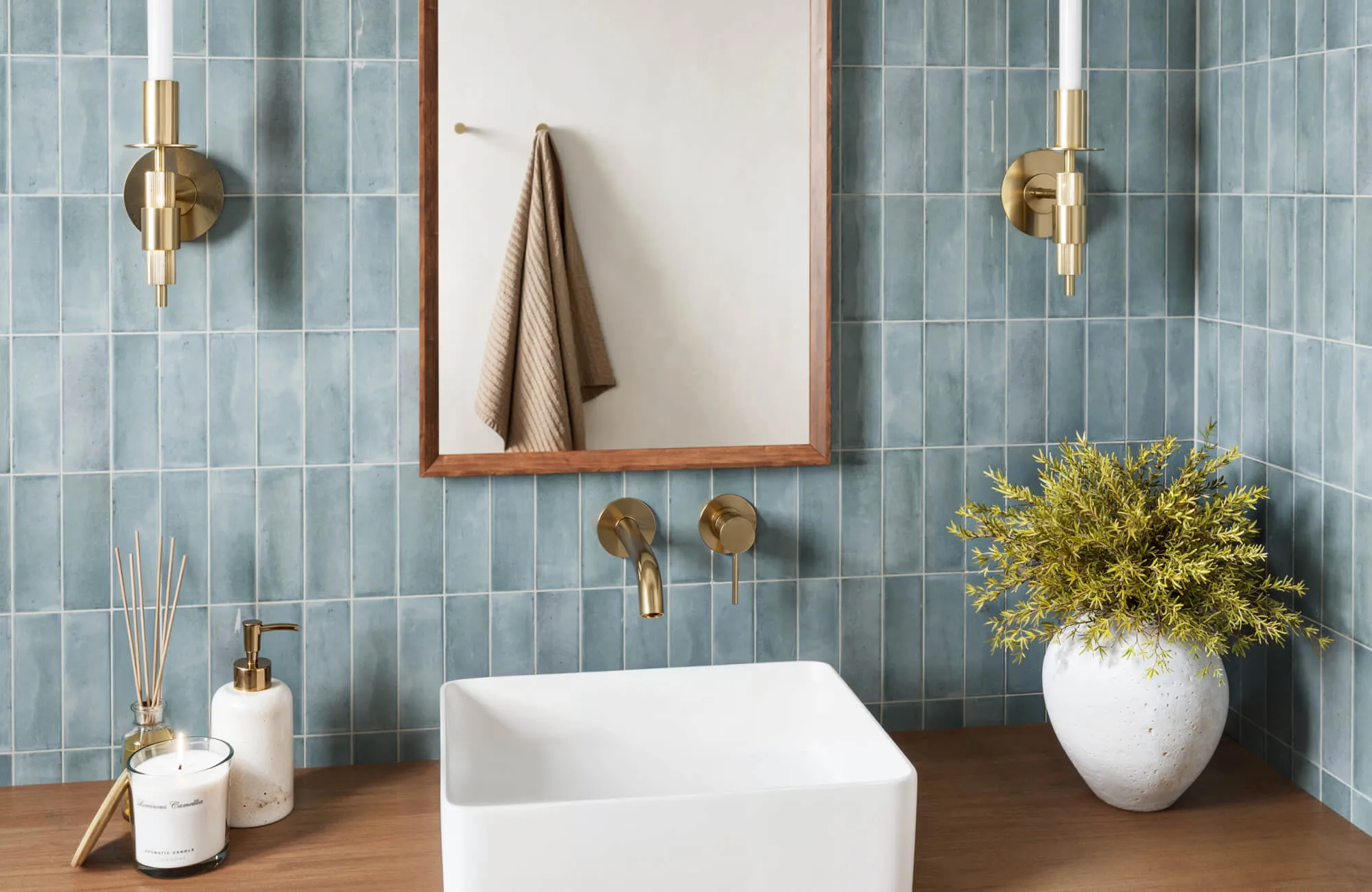 Soft blue subway tiles surround a bathroom vanity with brass lighting, a wood mirror frame, and a white sink on a warm wood counter.