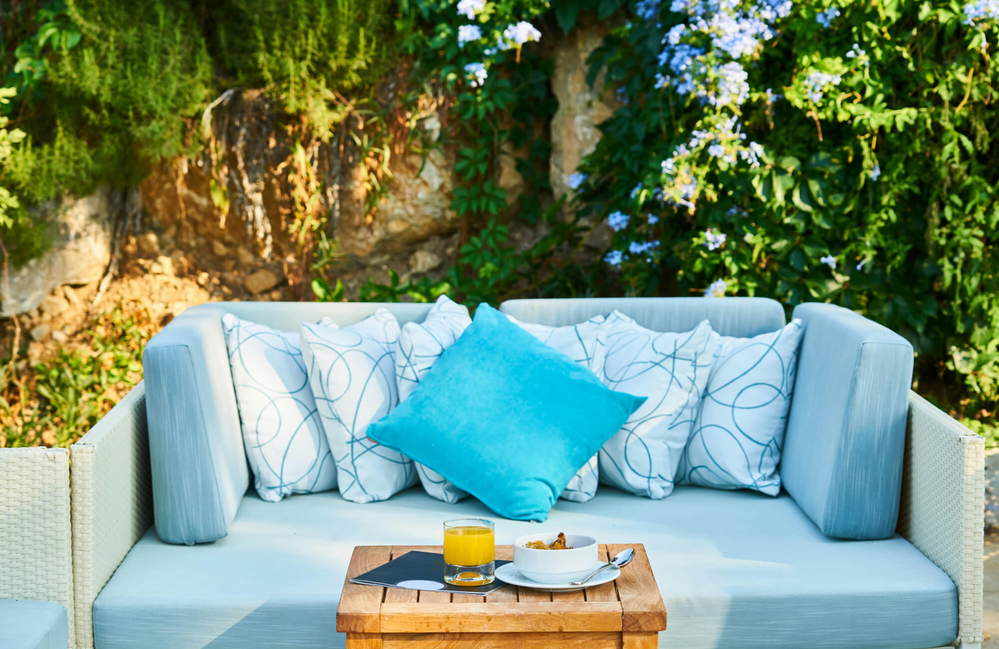 Outdoor wicker sofa styled with blue and white throw pillows, turquoise accent cushion, and wooden tray with juice and snacks.