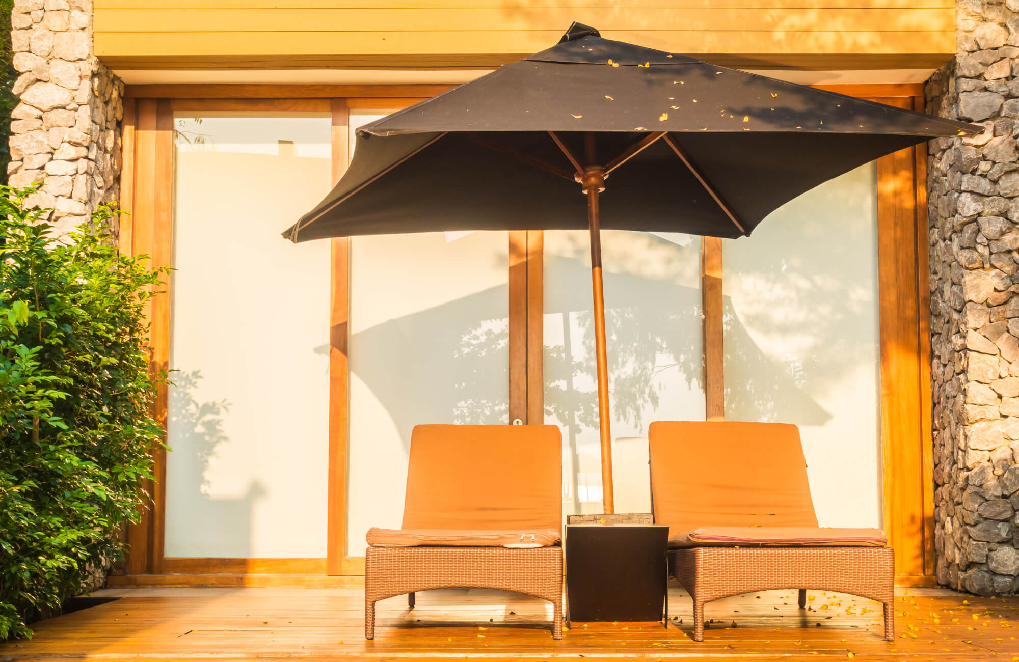 null Outdoor wicker loungers with warm orange cushions are styled beneath a black umbrella on a timber deck, highlighting a sun-protected seating area beside stone walls.