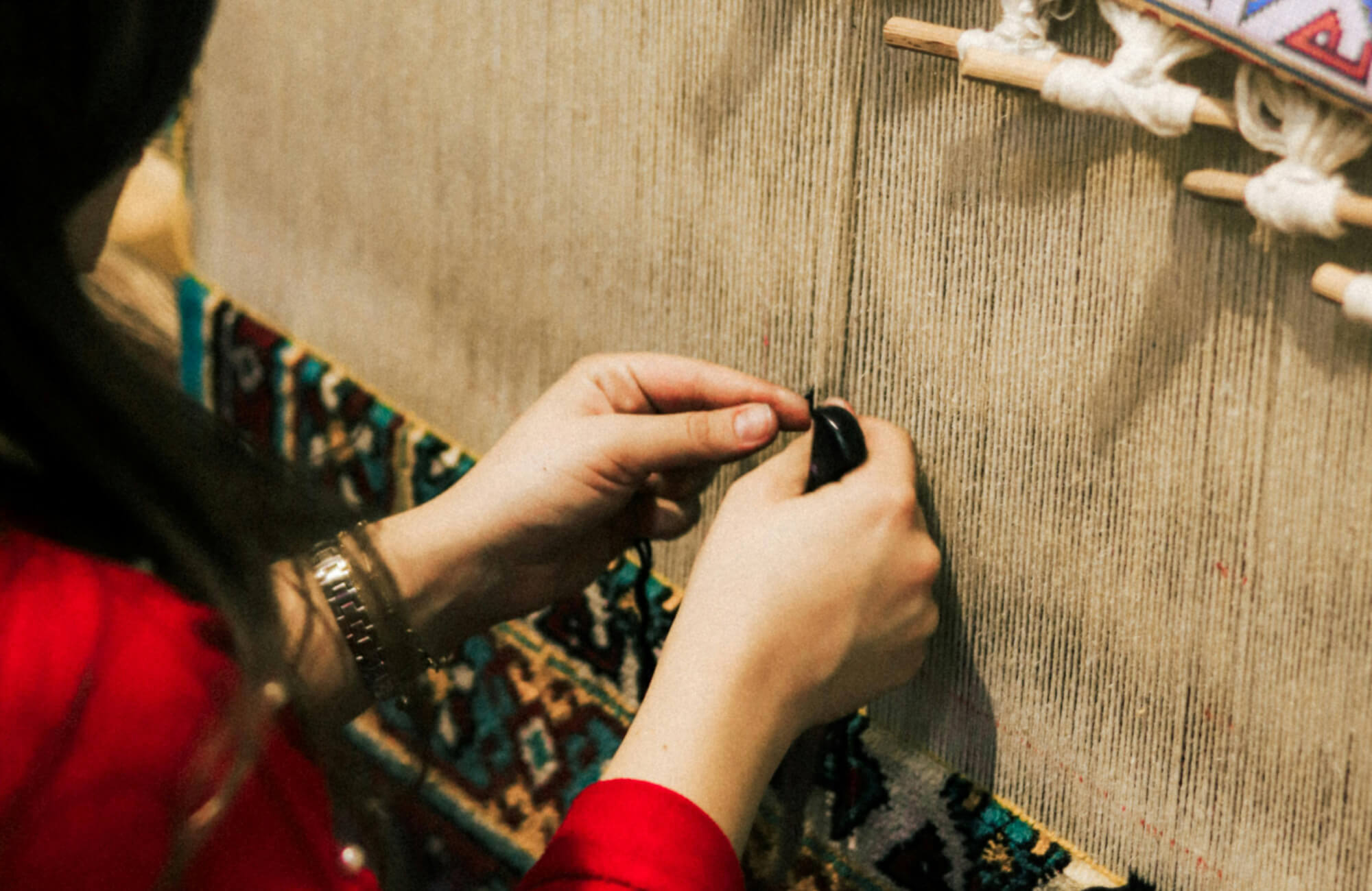 null Hands weaving an intricate multicolor rug pattern on a traditional loom, showcasing dense knots and fine textile construction.
