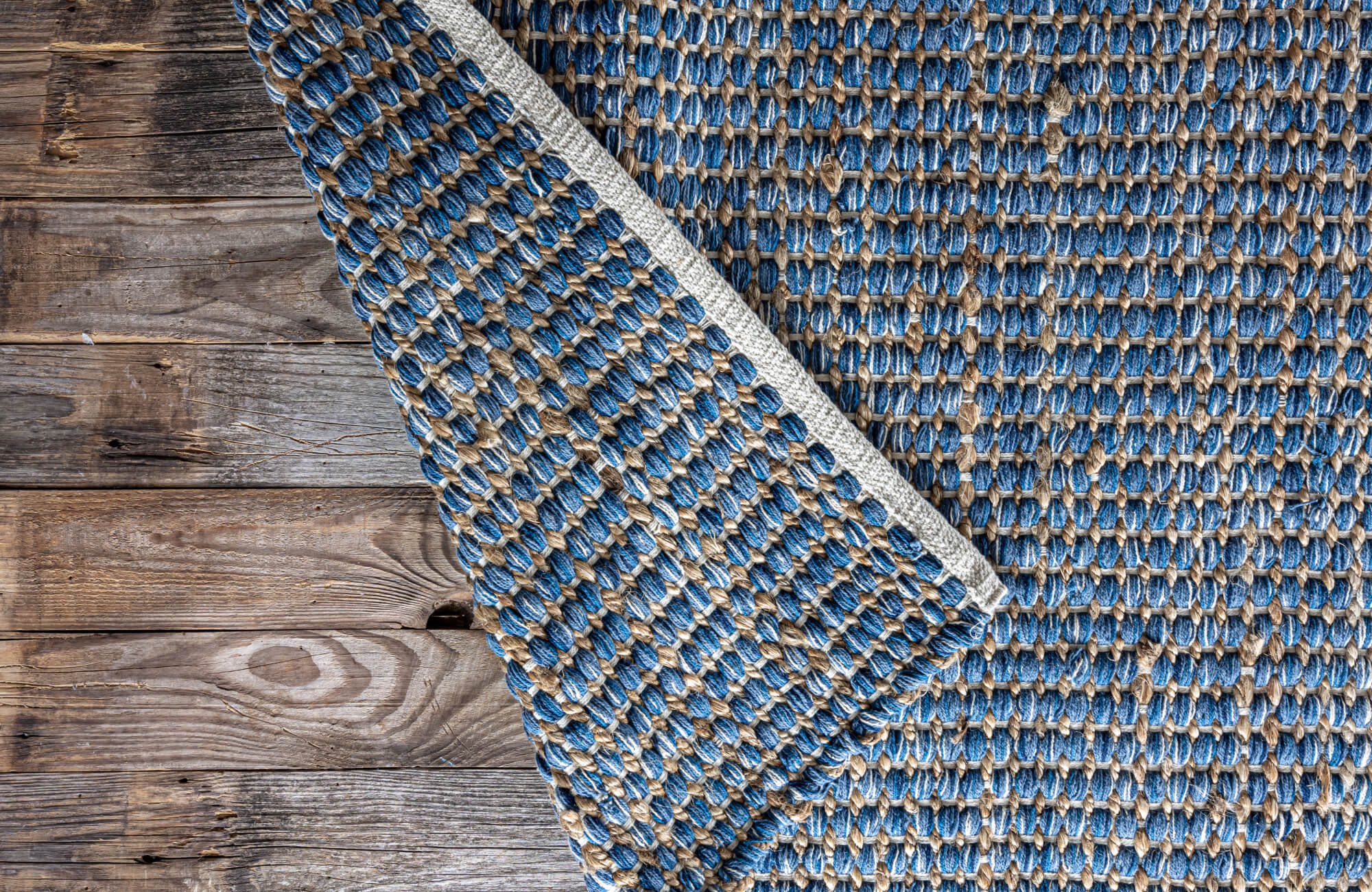 null Blue and tan basket-weave outdoor rug with visible backing detail, layered on weathered wood plank flooring.