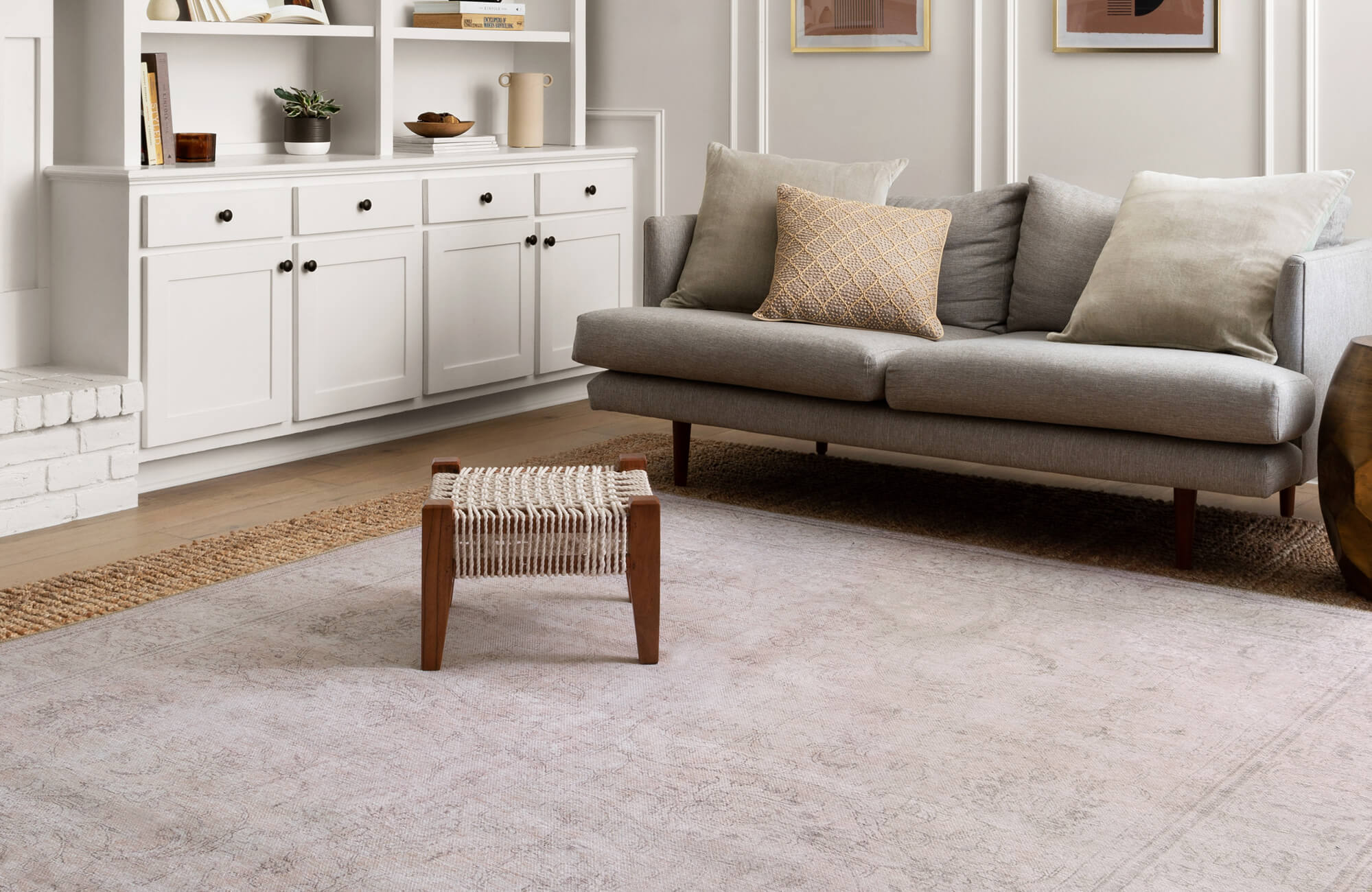 Bright, minimalist living room featuring a soft neutral rug, mid-century grey sofa, and woven wood stool on light oak flooring.