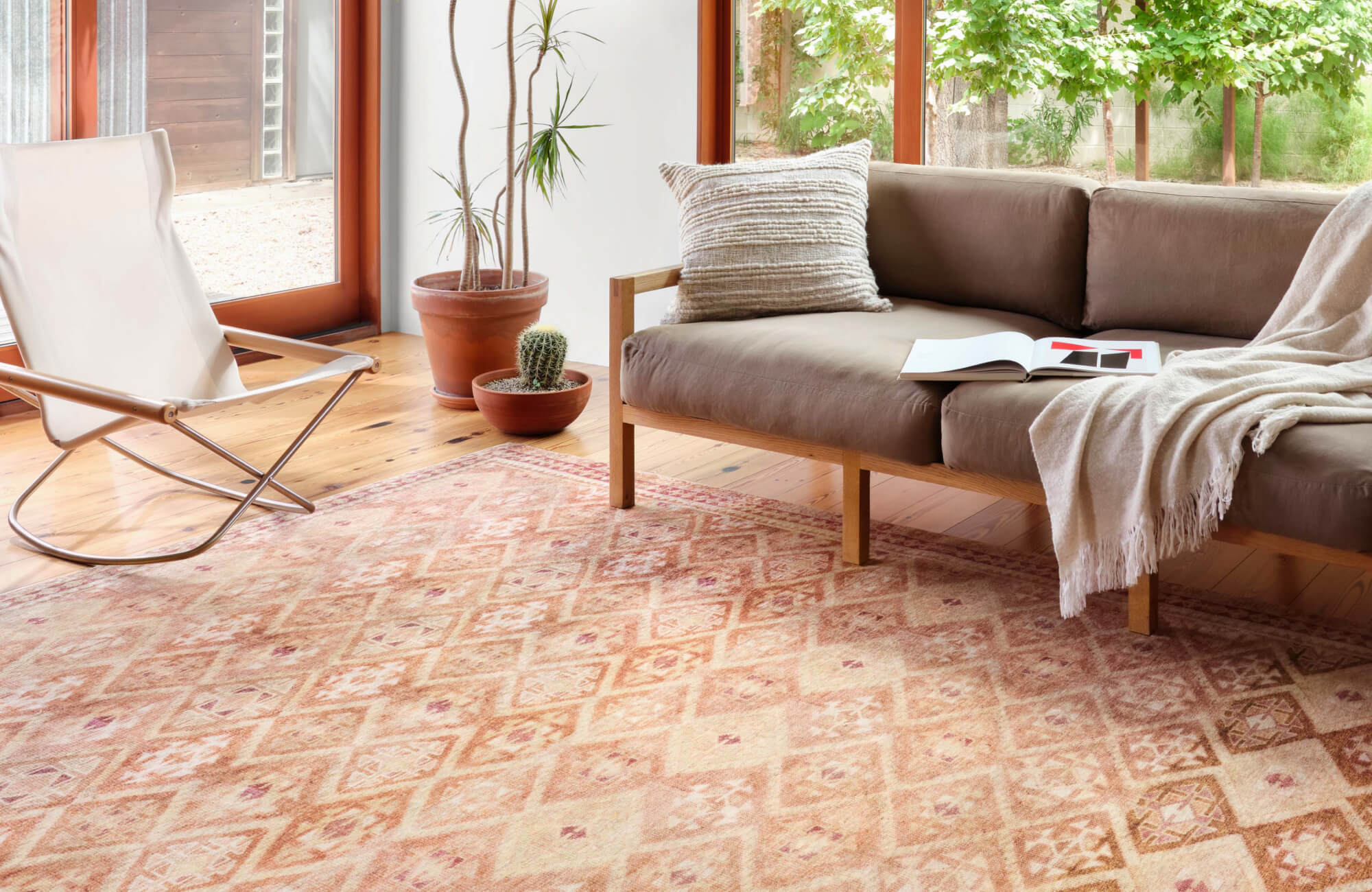 Sunlit living room with a terracotta-hued geometric rug, neutral wood-frame sofa, and desert-style potted plants.